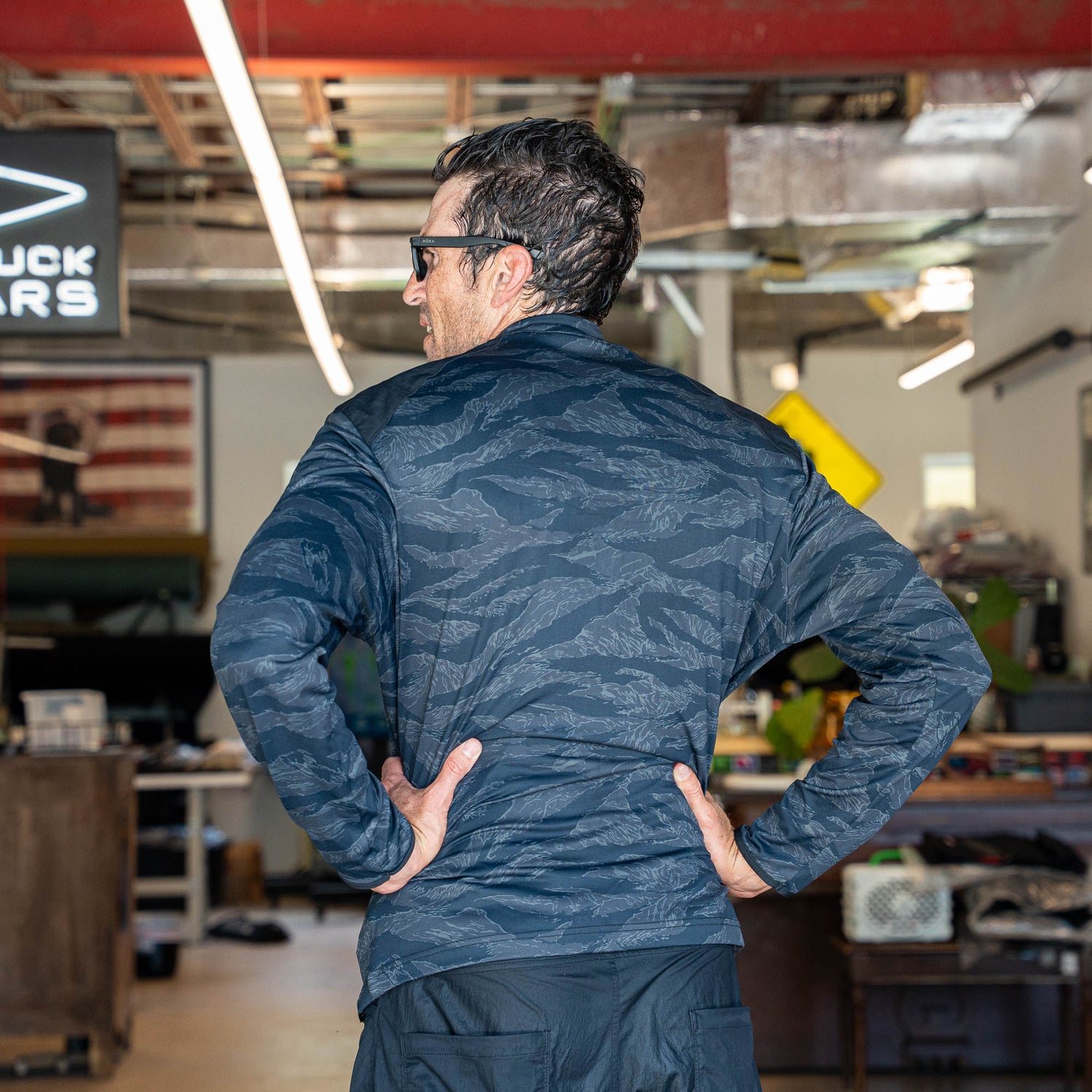 Man wearing sunglasses and a patterned, lightweight Warm Up Full Zip jacket with ToughDry shoulders stands indoors with hands on hips, facing away from camera. Made in USA.