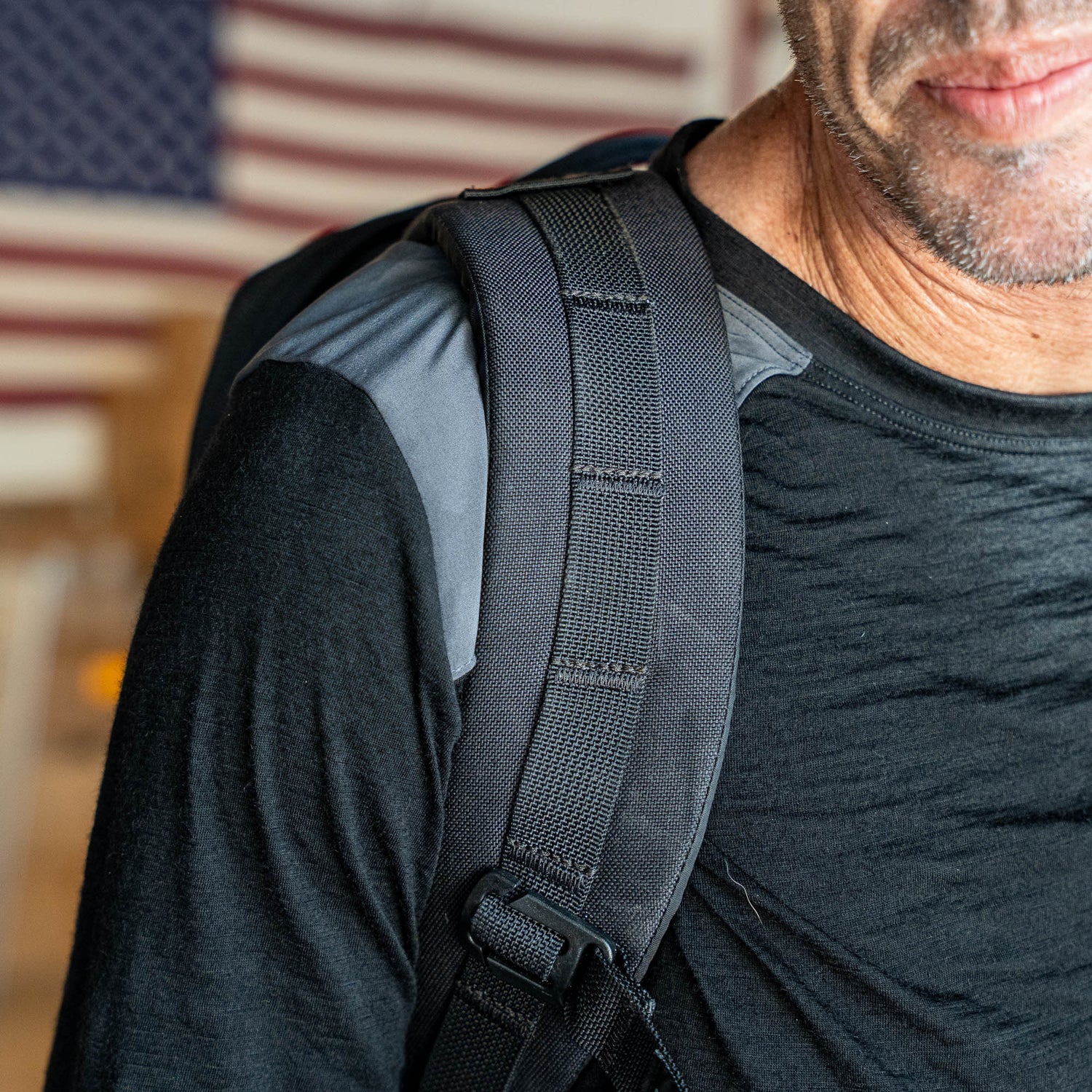 A man wearing the Men’s Commando Long Sleeve - Merino Wool shirt and a backpack stands in front of a blurred American flag.