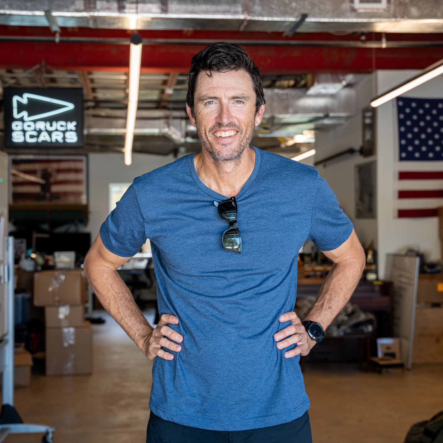 A man wearing the Slick Short Sleeve Tee - Tri-BlendX in blue stands smiling in a workshop, with American flags and boxes in the background.