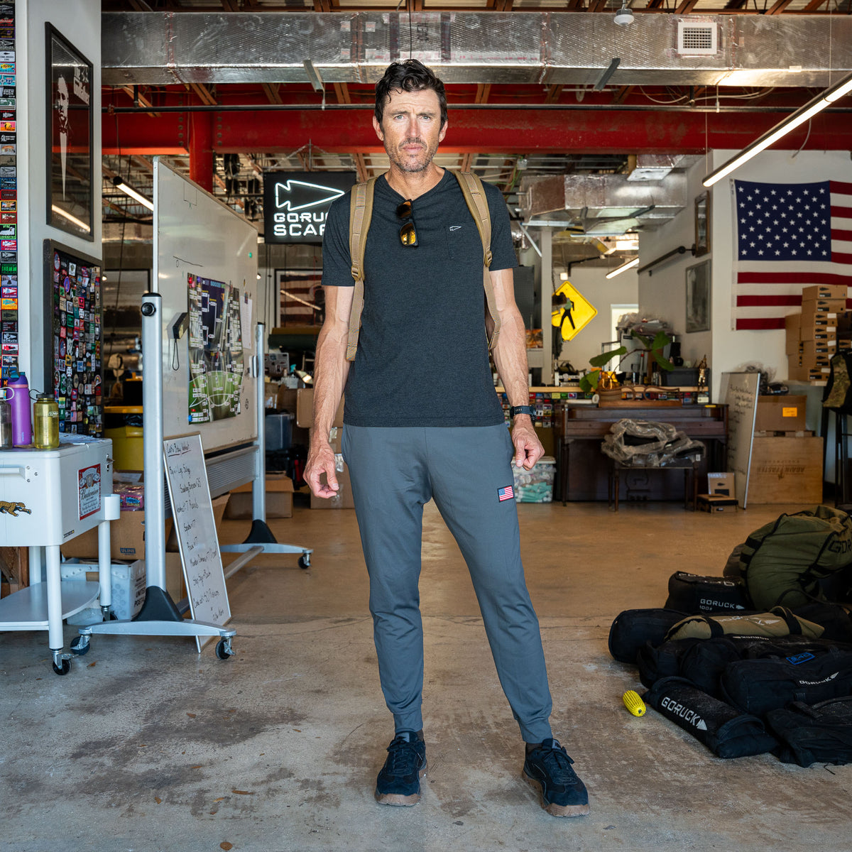 A man in workout clothes stands in a gym with an American flag and equipment in the background, wearing Men’s American Training Sweatpants—made in the USA and built to last with a lifetime guarantee.