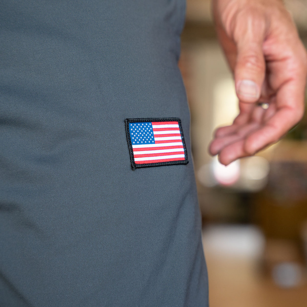A hand near a gray fabric with a small embroidered American flag patch highlights the Men’s American Training Sweatpants, proudly made in the USA for lasting durability.