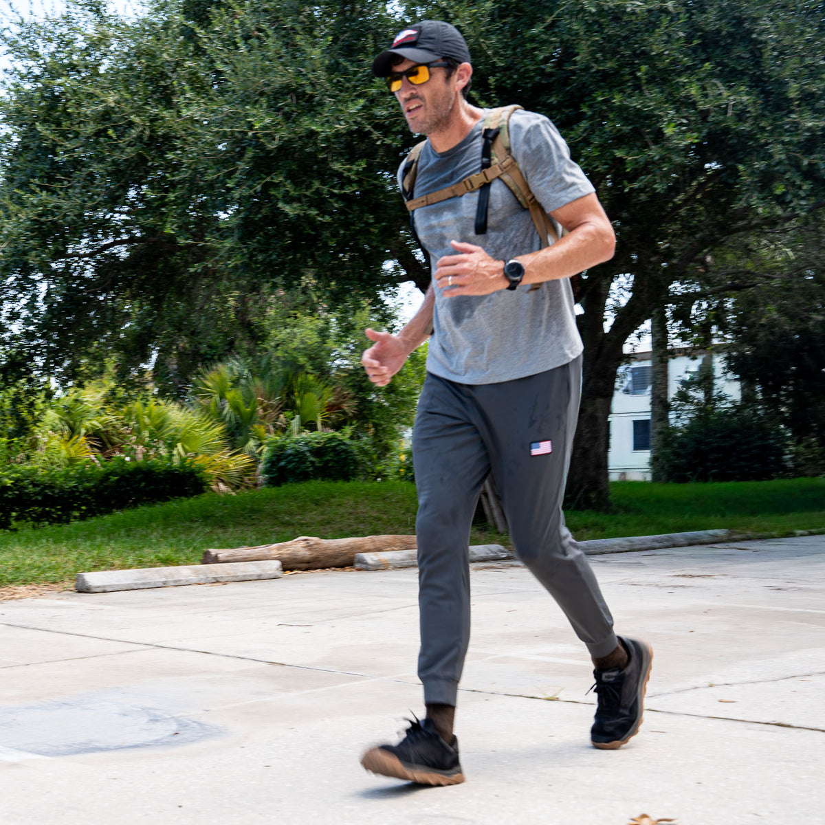 A man wearing a backpack jogs along a sidewalk in Men’s American Training Sweatpants, surrounded by trees and greenery on a sunny day.