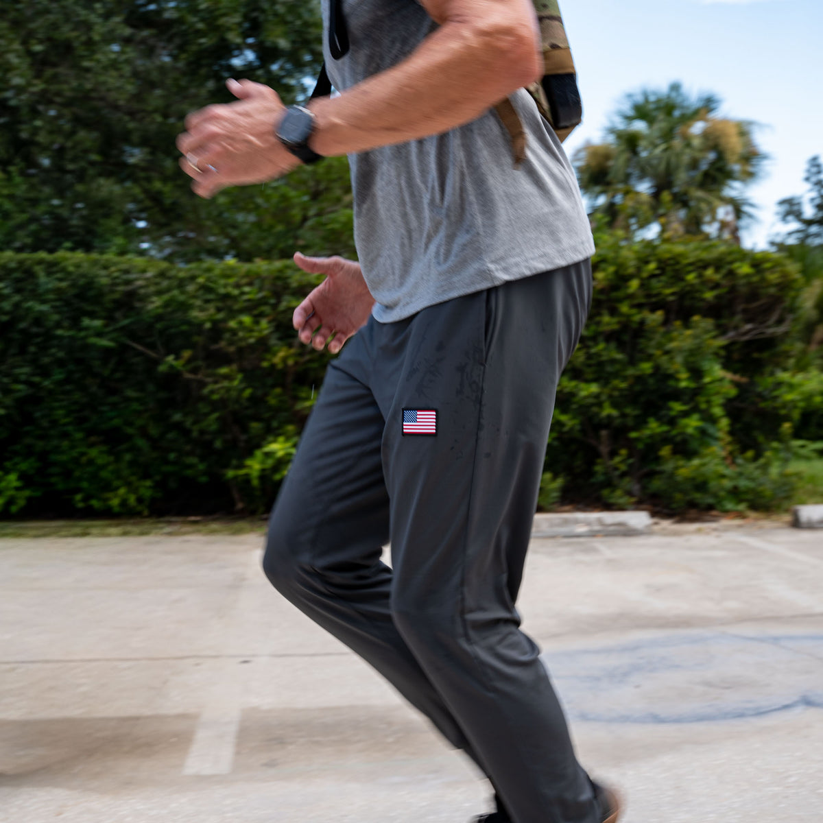 A person jogging outdoors in Men’s American Training Sweatpants with a US flag patch, paired with a gray shirt and smart watch. These durable sweatpants are made in the USA and ideal for active lifestyles.