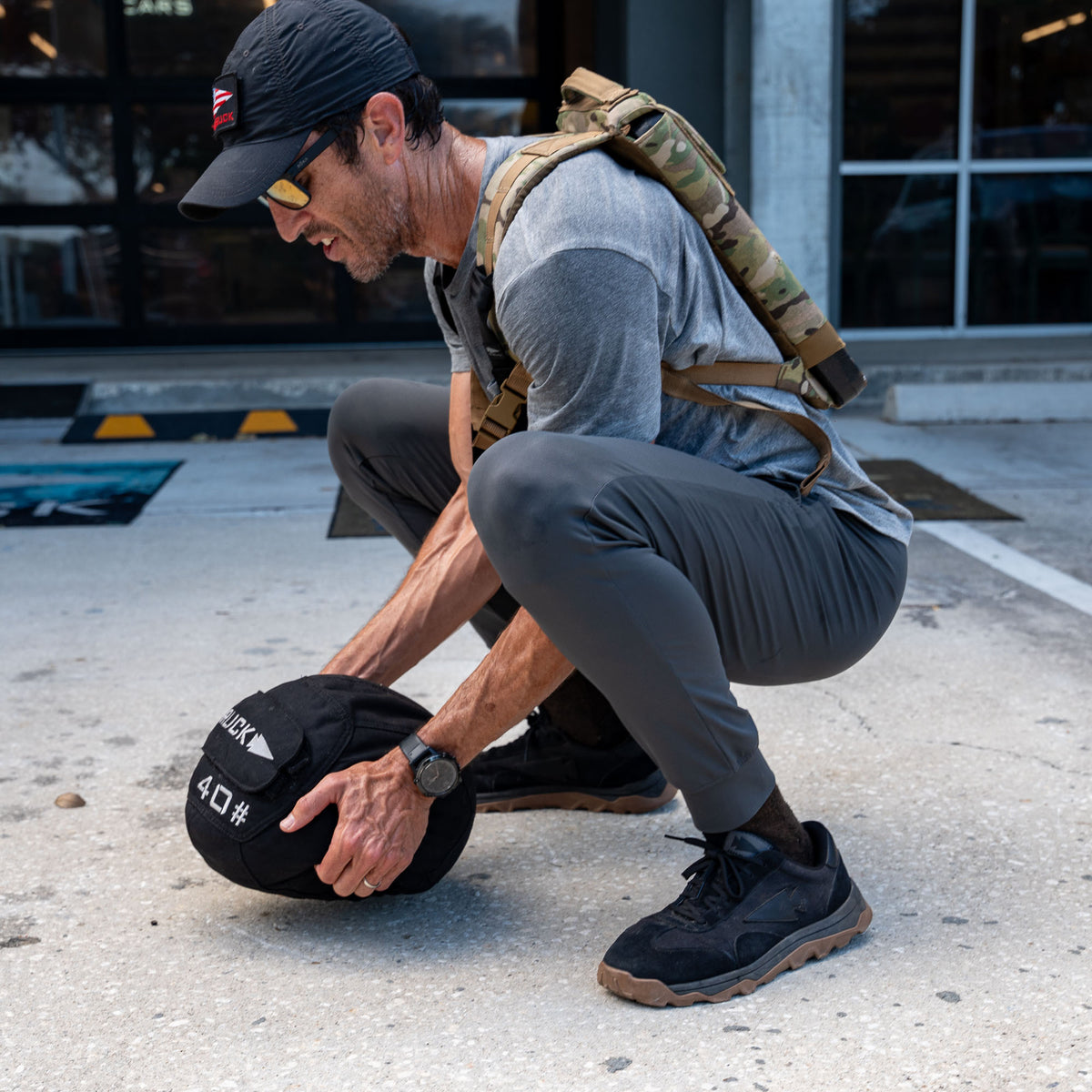 A man wearing Men’s American Training Sweatpants squats to lift a 40-pound sandbag outdoors, sporting a cap, sunglasses, and backpack.
