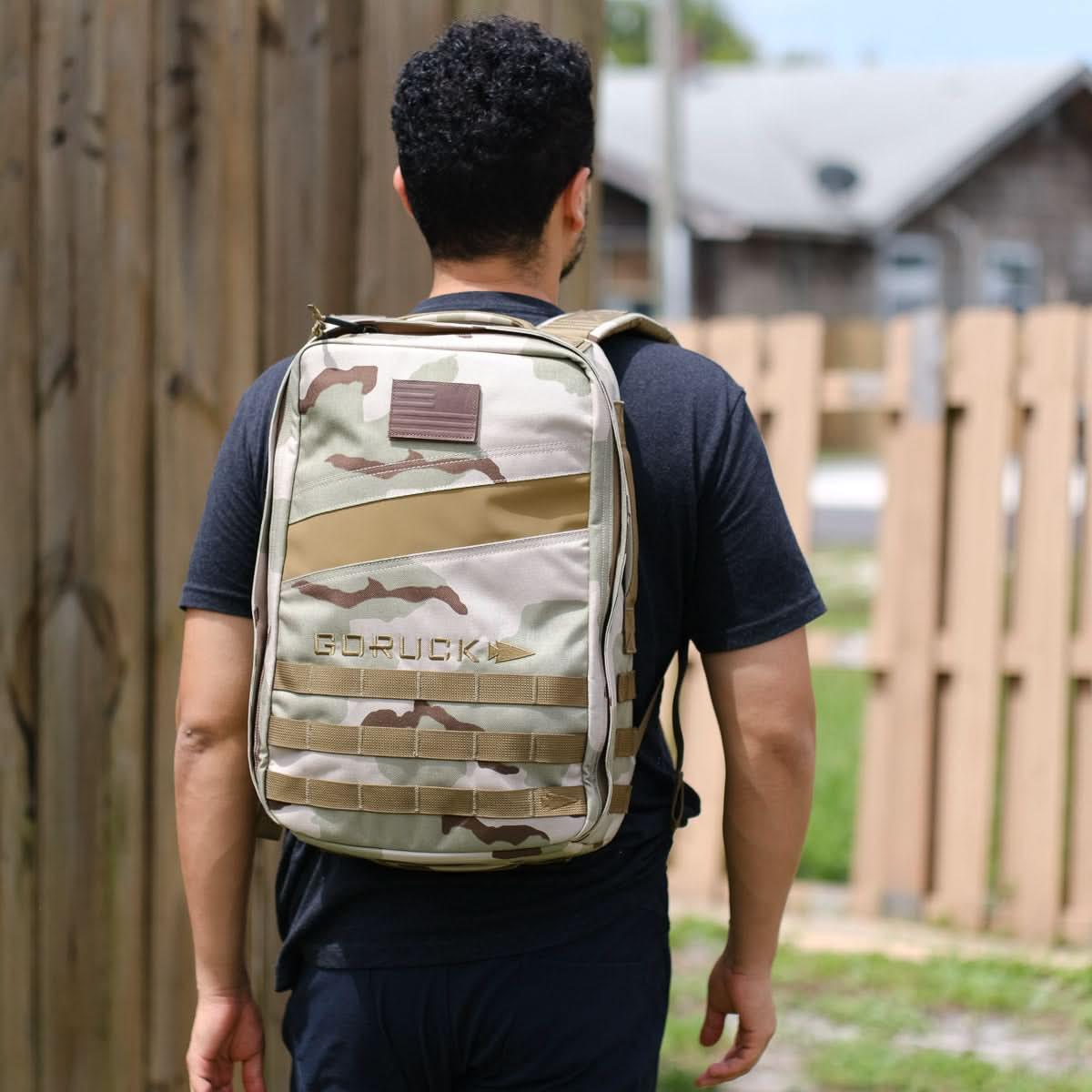 Man wearing a tan camouflage GORUCK backpack outdoors by wooden fence