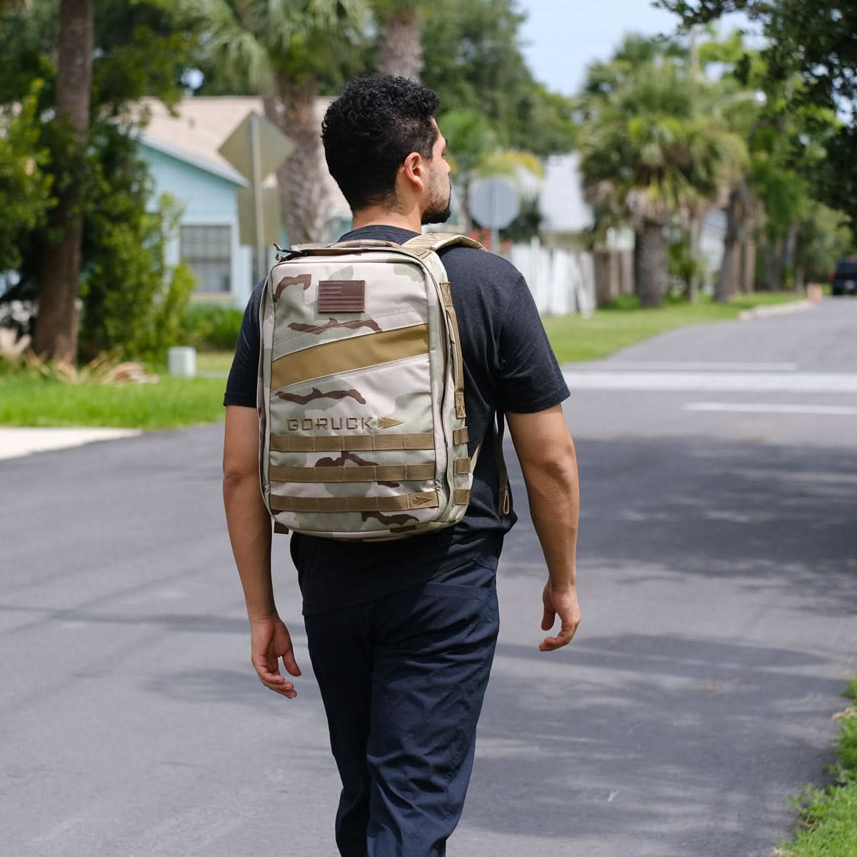 A man in dark clothing walks down a suburban street, rucking with the camouflage Rucker 4.0 backpack.
