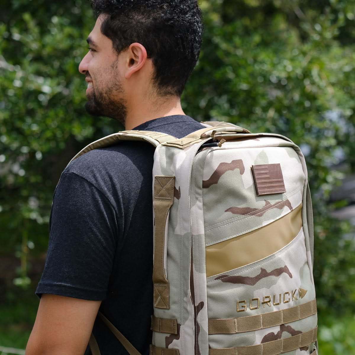 Man outdoors wearing a black t-shirt and a tan camouflage Rucker 4.0 rucksack with a patch on top.