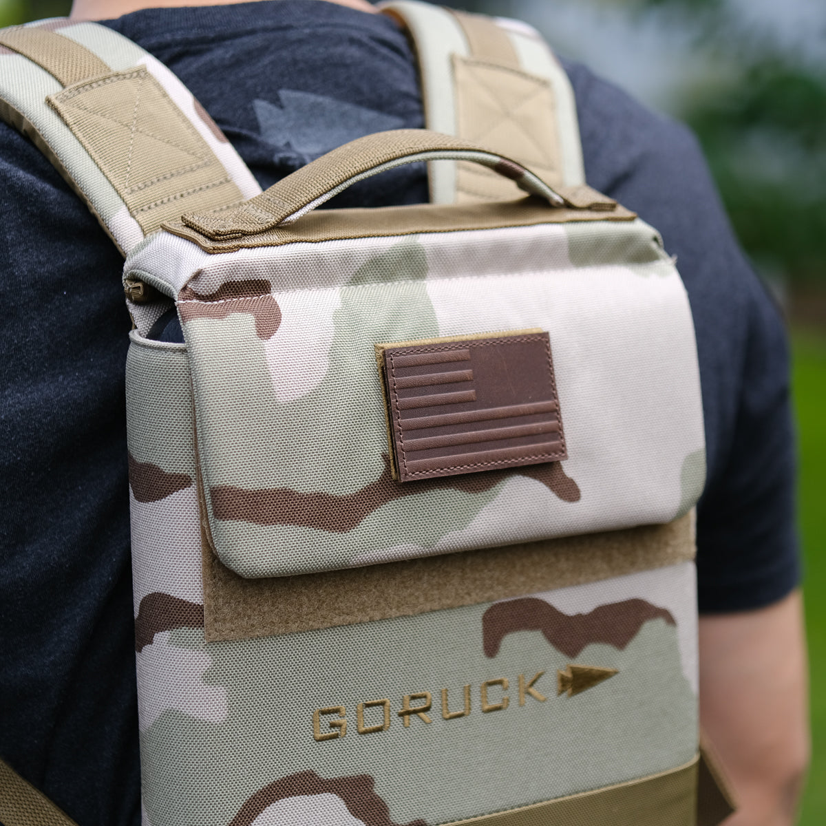 Close-up of a person wearing the Ruck Plate Carrier 3.0, a camouflage GORUCK backpack made from rugged ballistic nylon, featuring a subdued American flag patch.