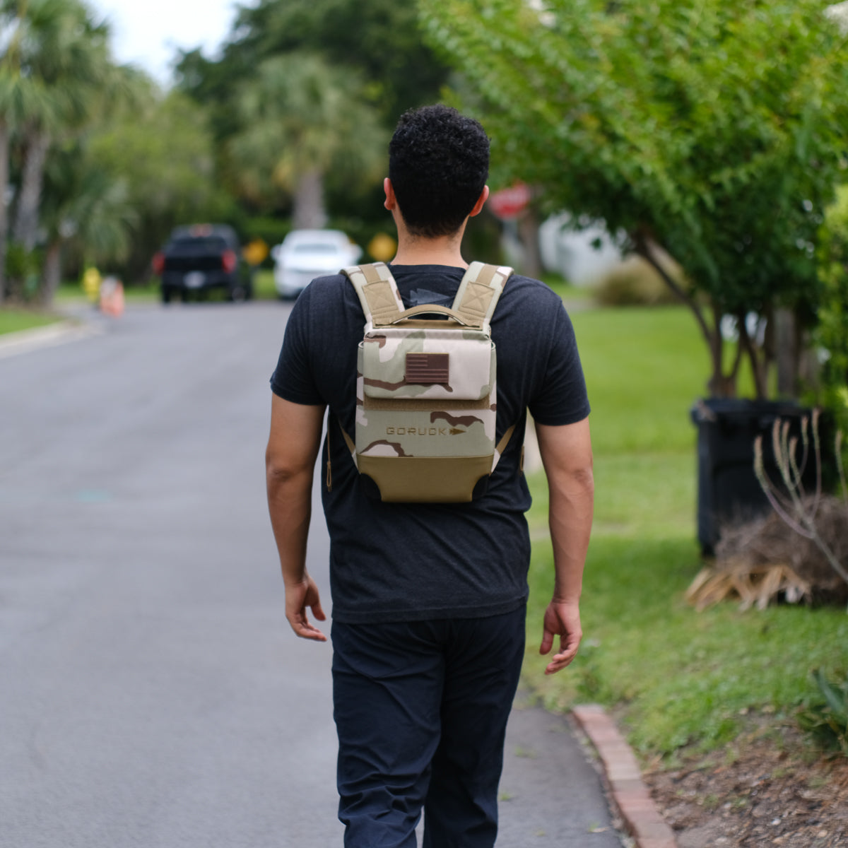 A man seen from behind walking on a suburban street wears black clothes and the Ruck Plate Carrier 3.0, a camo backpack made of ballistic nylon.