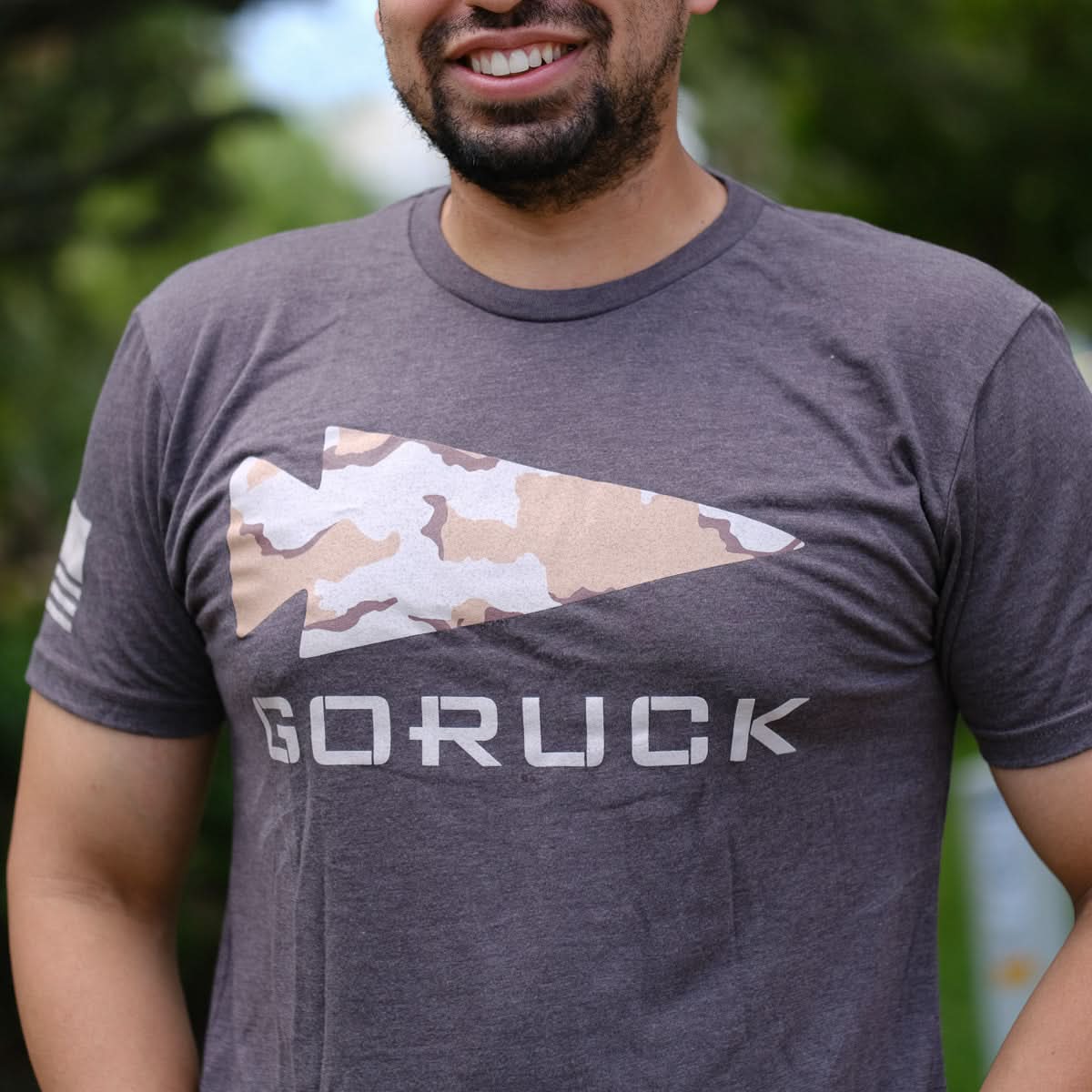 Smiling man wearing a GORUCK Logo DCU Camo Tee - Tri-Blend, featuring a camo spearhead design, standing outdoors.