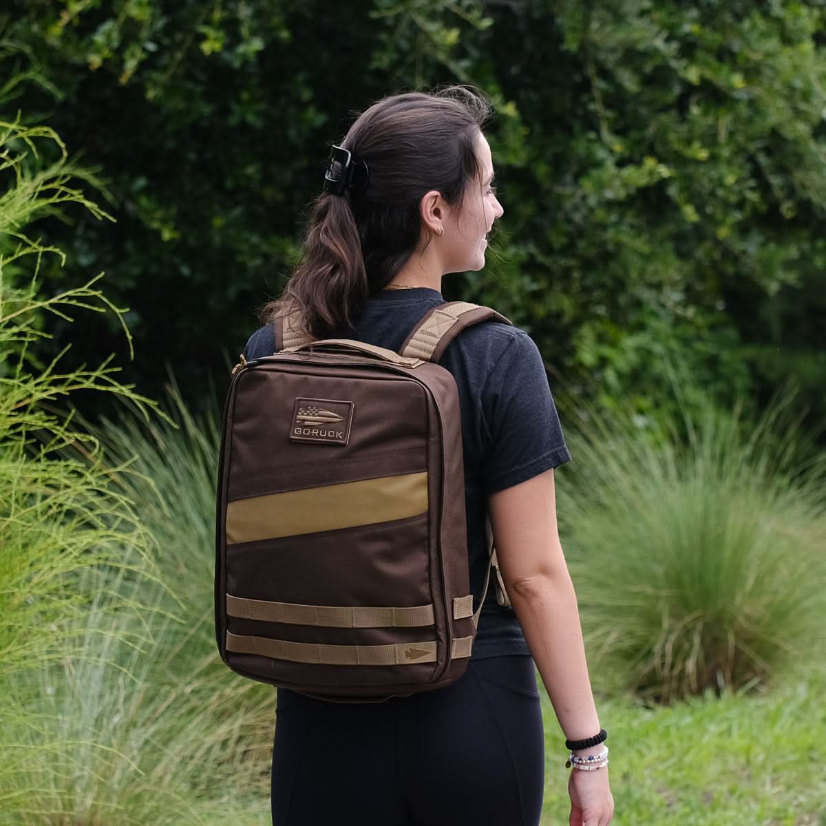 A woman with a brown Rucker 4.0 rucksack stands outside, surrounded by greenery—ideal for rucking or carrying your favorite ruck plates.
