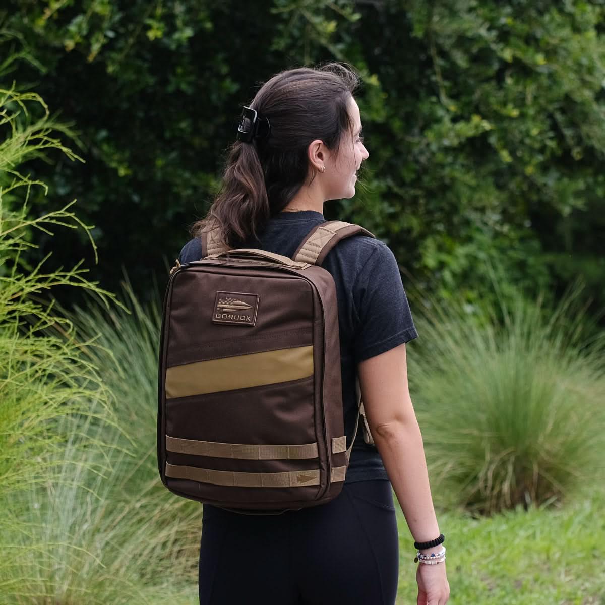 Woman wearing brown GORUCK backpack outdoors with greenery background