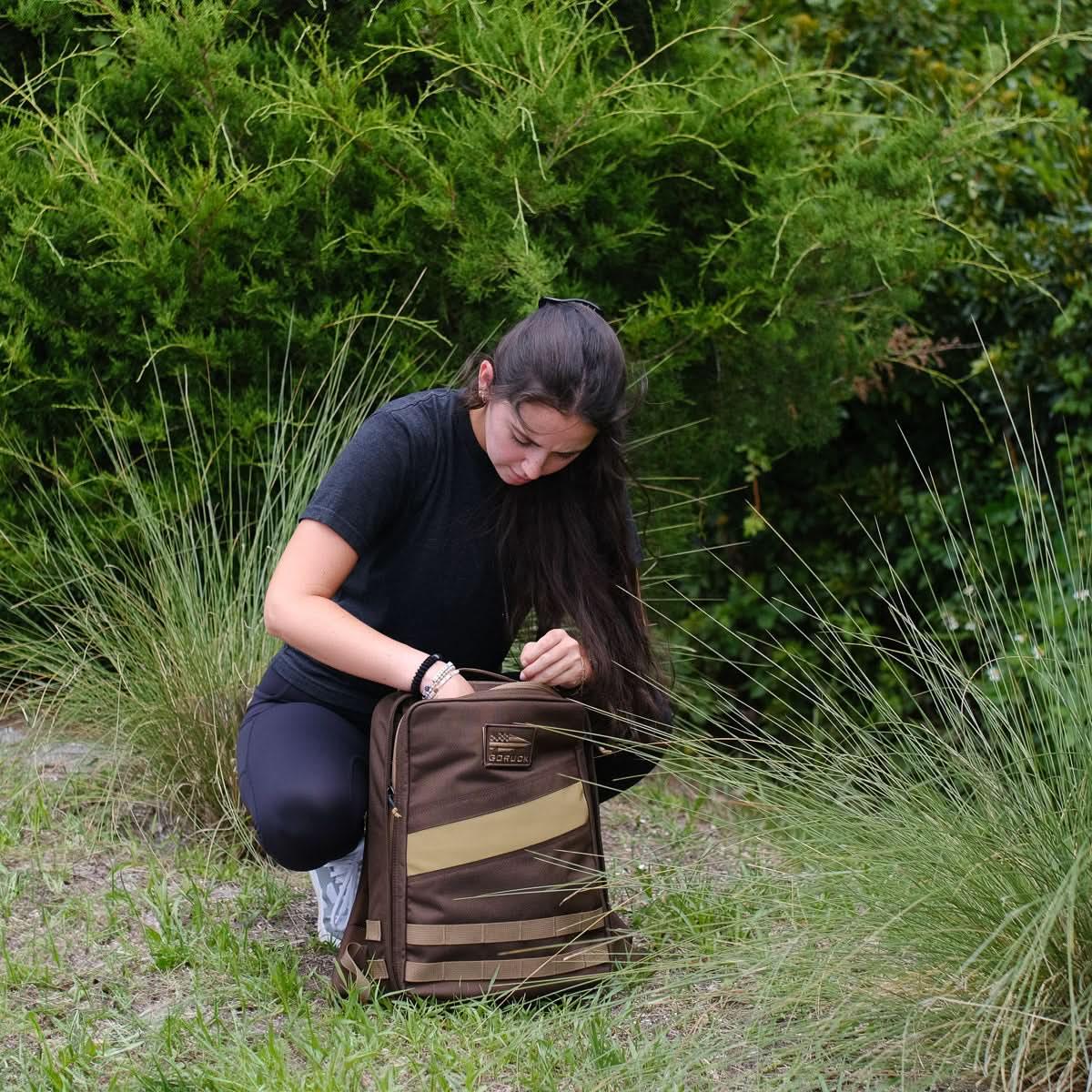 Woman outdoors kneeling in grass opening a brown GORUCK rucksack with green shrub background