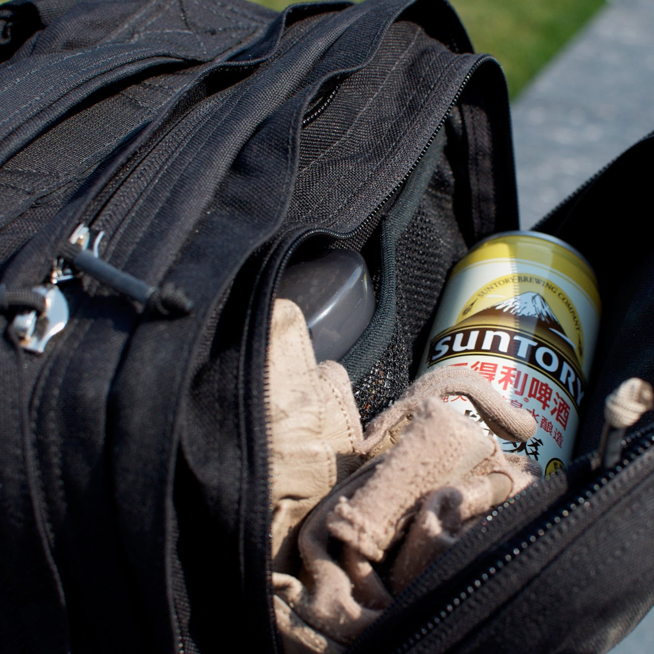 A Suntory drink can, gloves, and a case rest inside the open pocket of a black GR2 USA - Cordura carry-on backpack outdoors.