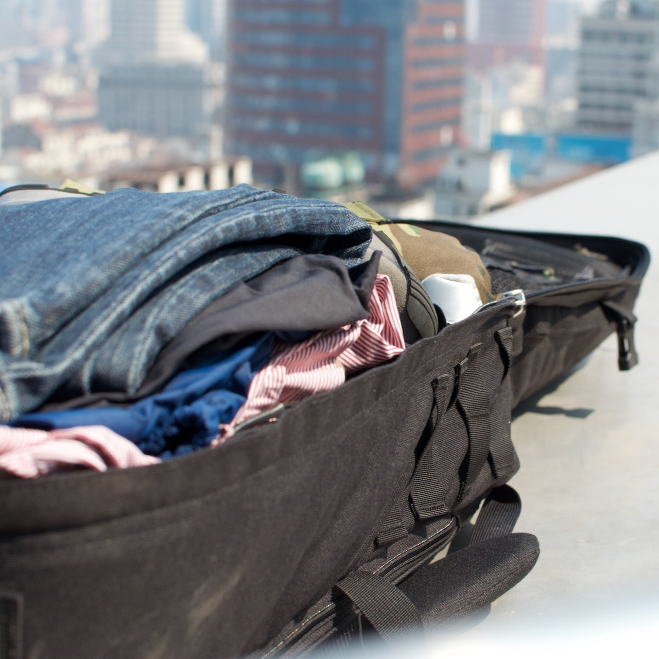 The GR2 USA - Cordura carry-on backpack, packed with clothes, sits open on a rooftop with city buildings in the background.