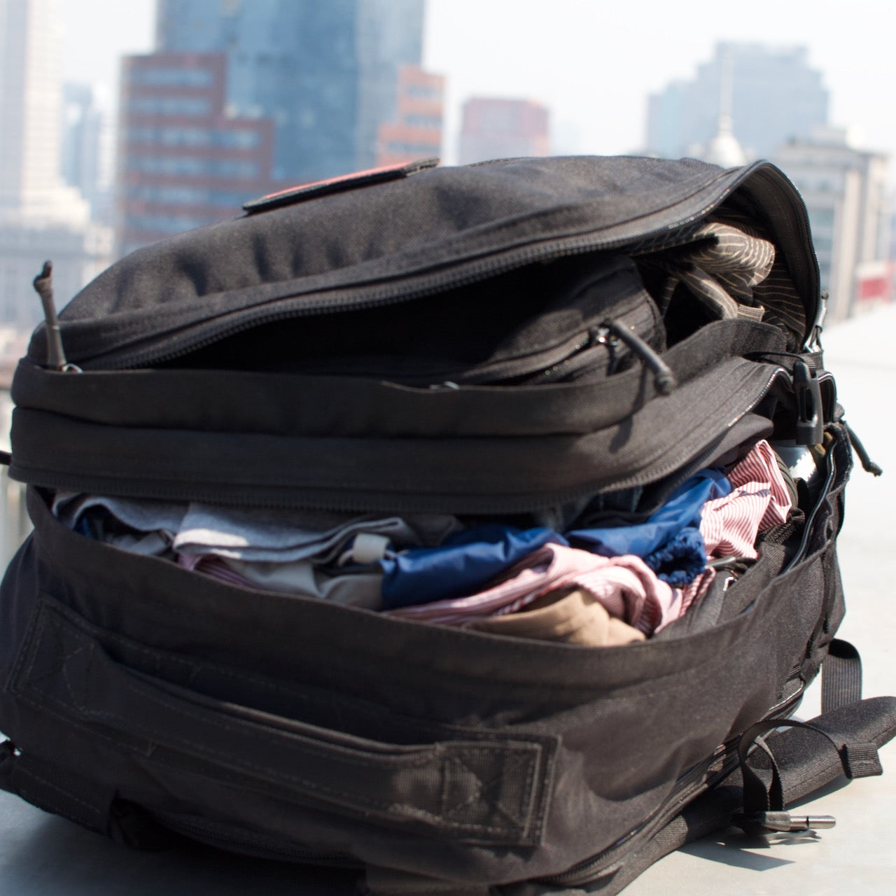 The GR2 USA - Cordura black travel backpack, packed with clothes, sits open on a rooftop with city buildings in the background.