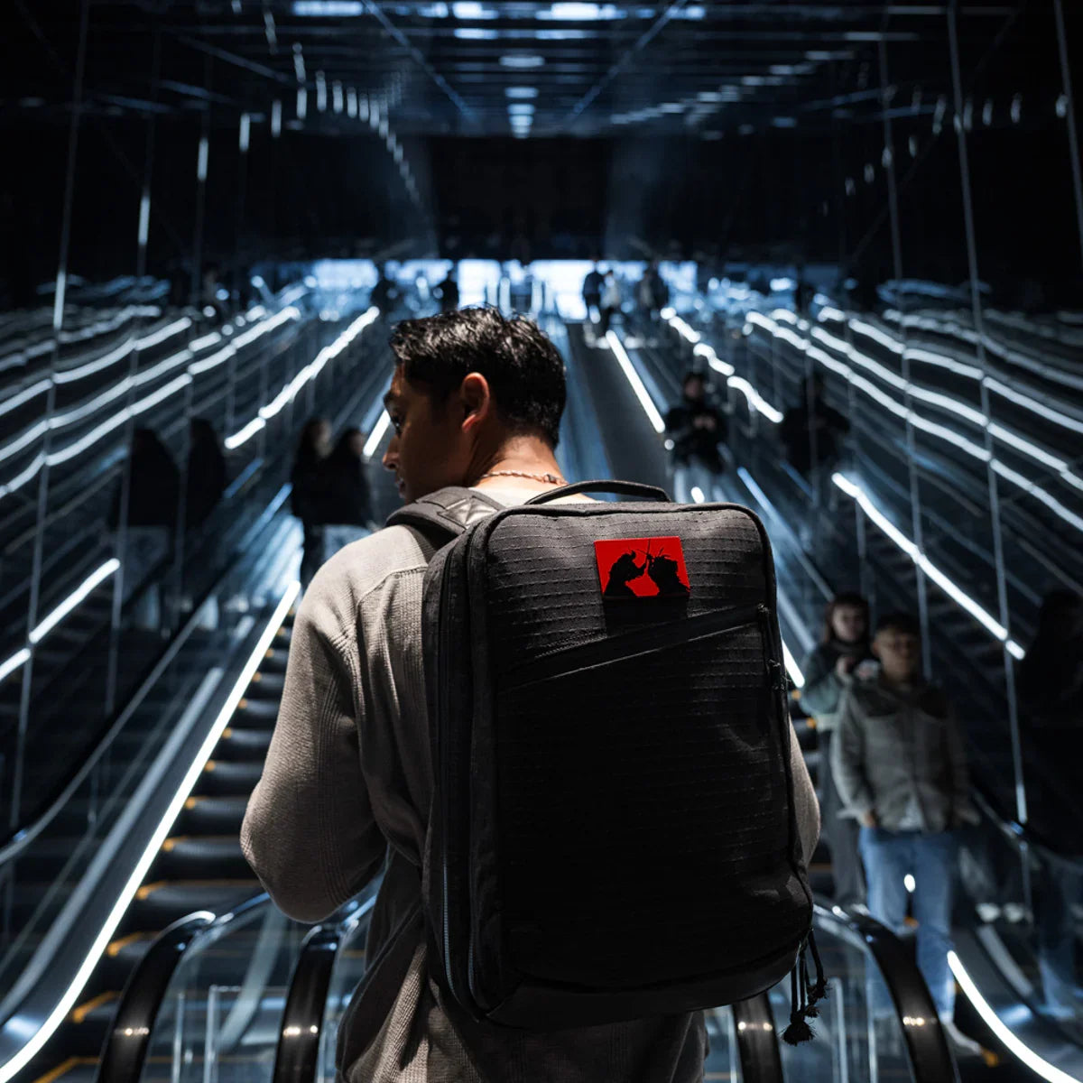 Man carrying black GORUCK backpack with red patch on escalator in dimly lit modern urban setting