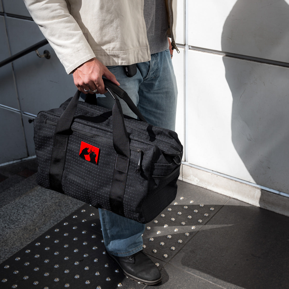 A person in jeans and a jacket stands outside holding the Kit Bag x Carryology - GRXC2 Samurai 2.0, which is crafted from custom technical fabric and features a red and black logo.
