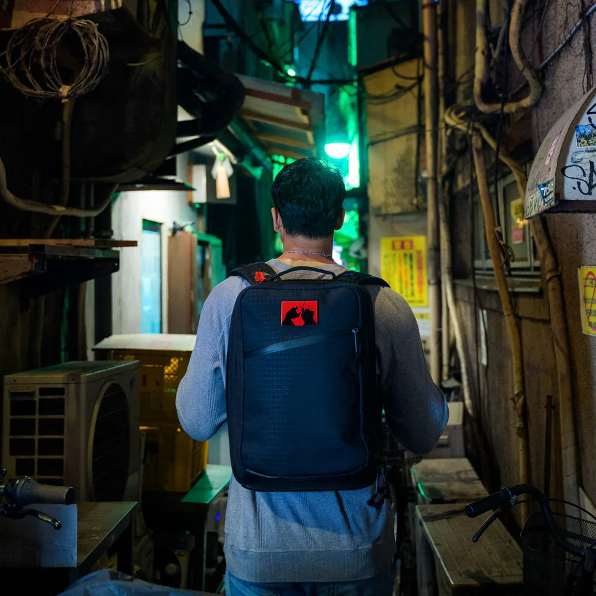 Man wearing GORUCK backpack walking through narrow, dimly lit urban alley with neon lights