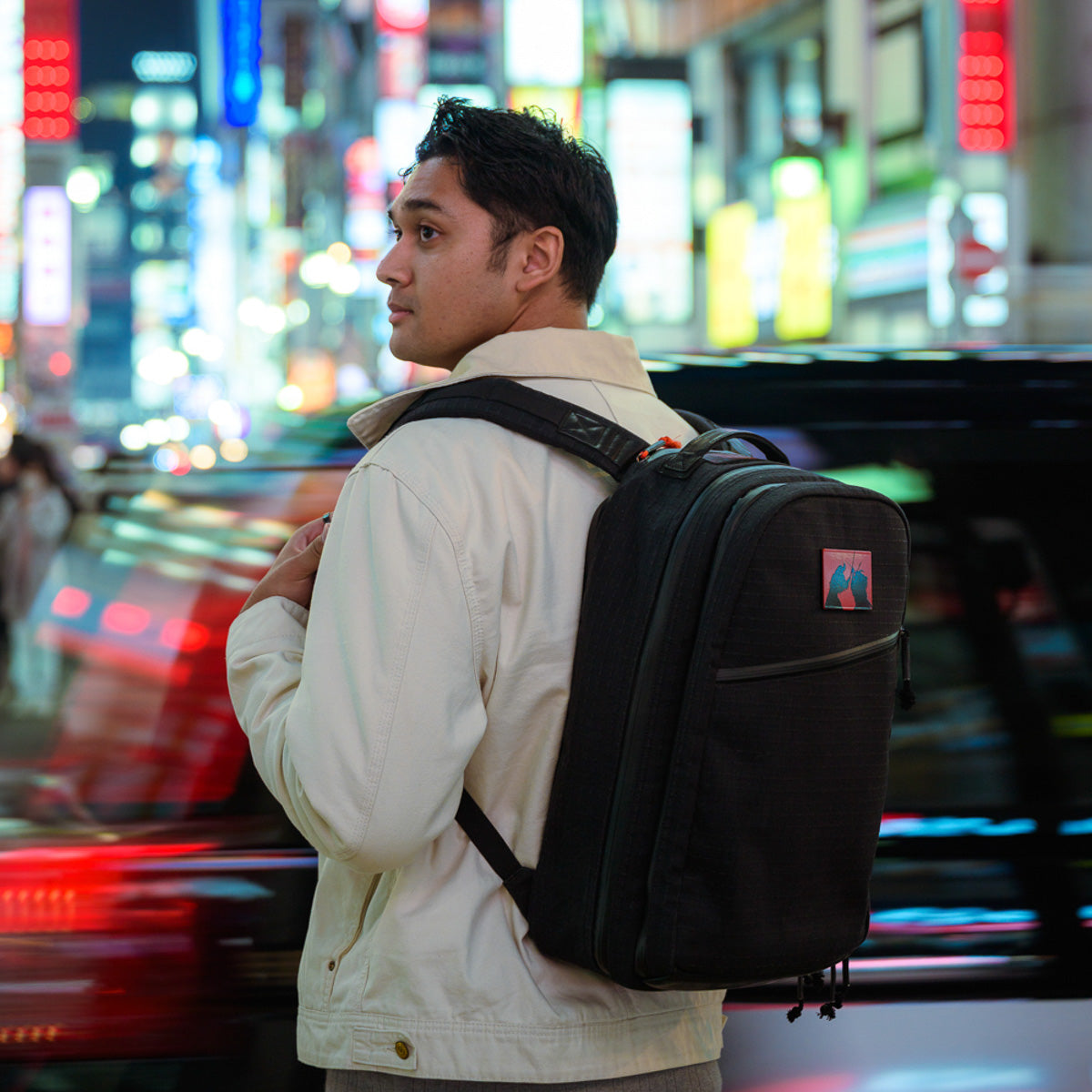 A man with a Bullet Ruck x Carryology - GRXC2 Samurai 2.0 backpack stands on a bustling city street at night, illuminated by vibrant lights and passing cars.