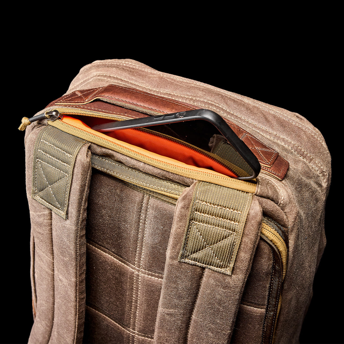 The GR1 USA - Heritage Waxed Canvas rucksack, featuring an orange lining, is shown unzipped with a black tablet inside against a black background.