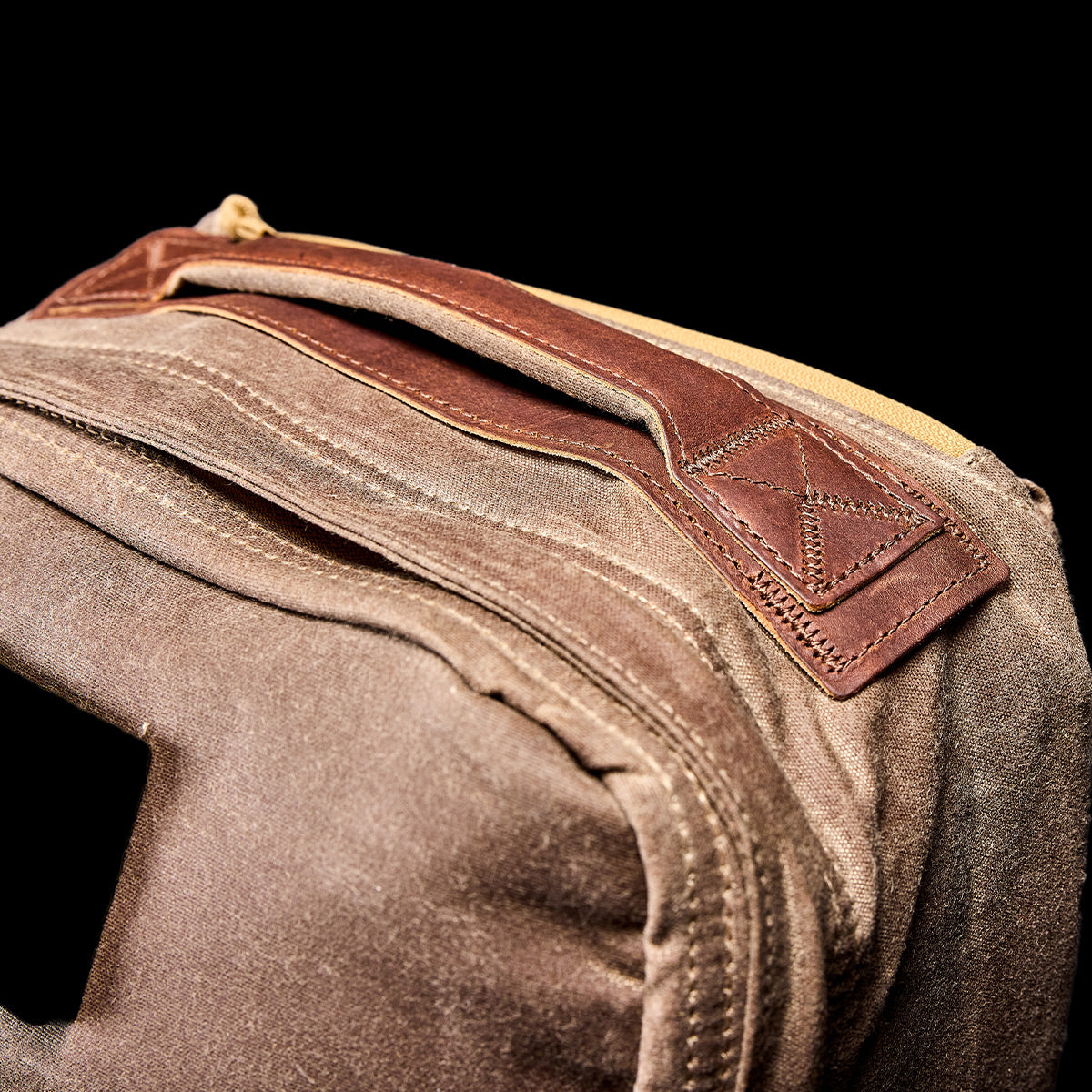 Close-up of the GR1 USA - Heritage Waxed Canvas rucksack, made from waxed duck canvas with a leather handle and yellow zipper, shown against a black background.