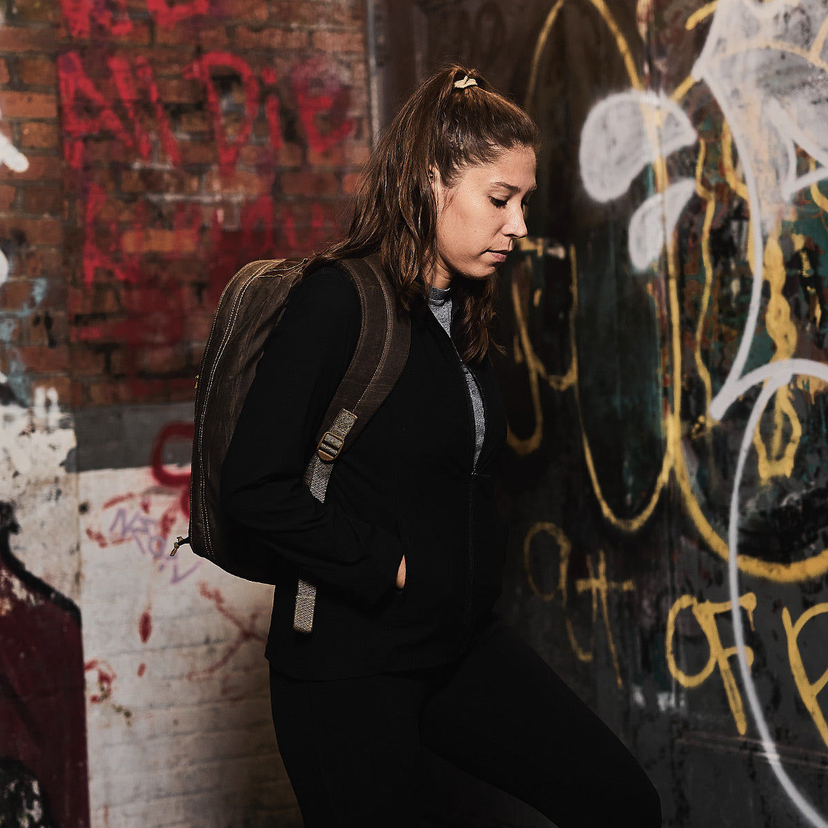 A woman with a GR1 USA - Heritage Waxed Canvas rucksack stands by a graffiti-covered wall, looking down with her hands in her pockets.