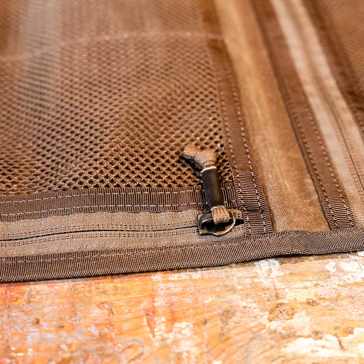 Close-up of the Wire Dopp - Waxed Canvas by GORUCK, featuring a brown mesh fabric with a zipper and toggle fastening on a wooden surface, highlighting its intricate stitching. Perfect for travel essentials, the textured design ensures optimal organization while providing durability similar to waxed canvas.