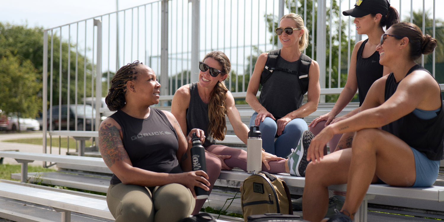 Five women in athletic wear sit and chat on bleachers outdoors, smiling and holding water bottles.