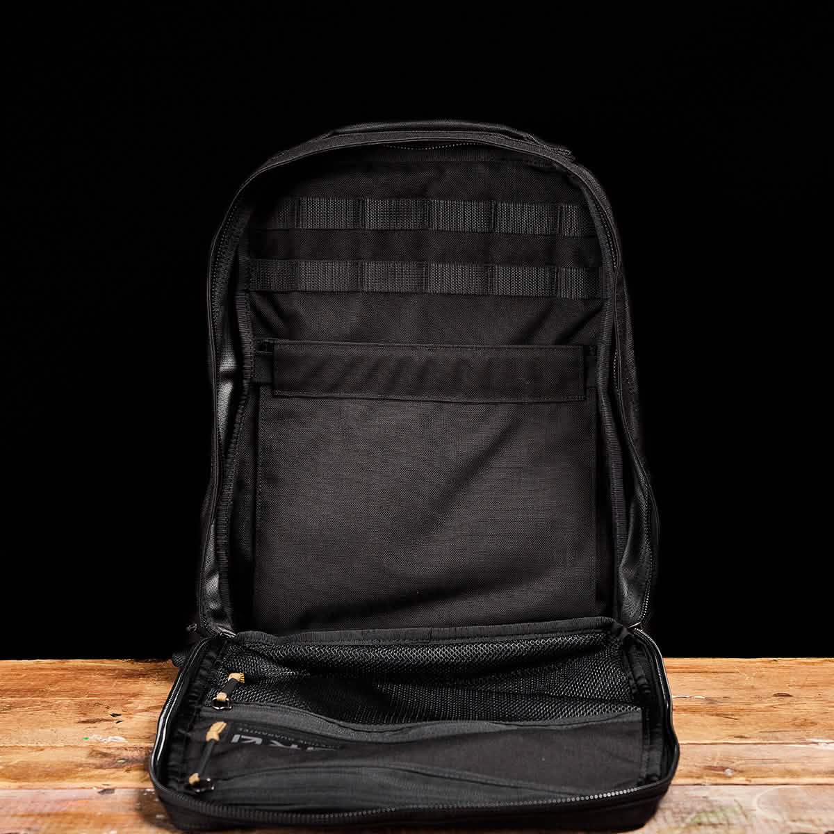 A sleek black GR0 - Cordura backpack by GORUCK sits open on a wooden surface, highlighting its minimalist EDC design. Against a black background, the interior displays mesh pockets and sections for organizing essentials, including a bombproof laptop compartment.
