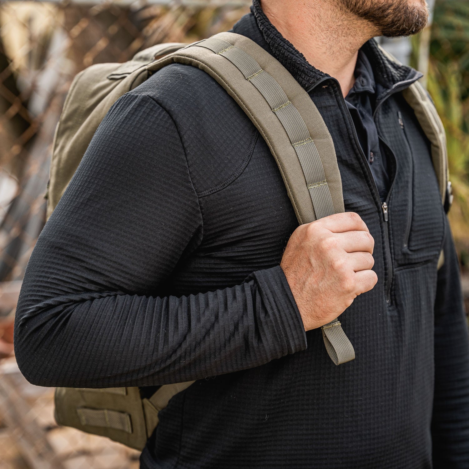Man in a black long-sleeve shirt carries a khaki GR1 USA - Cordura rucksack, an everyday carry backpack made to Special Forces standards, outdoors, shown from the shoulders up.