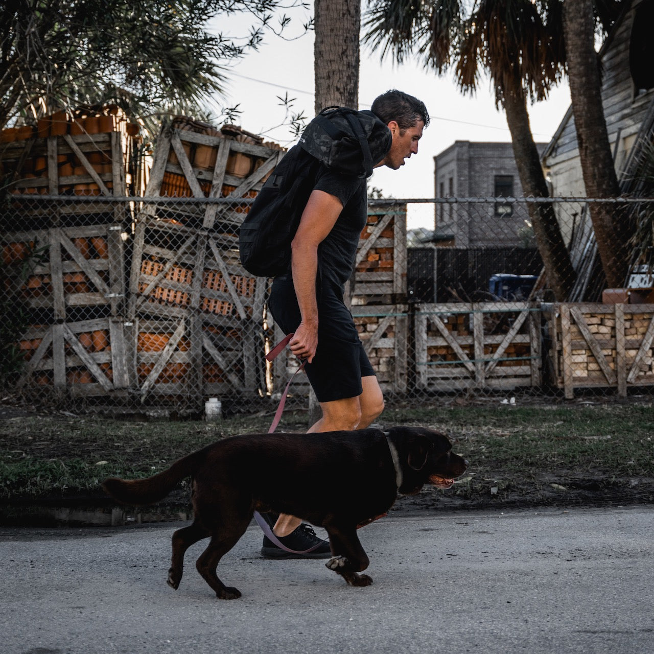 Man wearing Men’s Simple Shorts - Lightweight ToughDry® walks a brown dog on a leash along a street lined with wooden crates in the background.