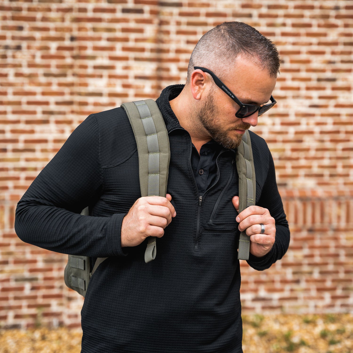 Man wearing sunglasses and the GR1 USA - Cordura everyday carry backpack stands against a brick wall, looking down thoughtfully.