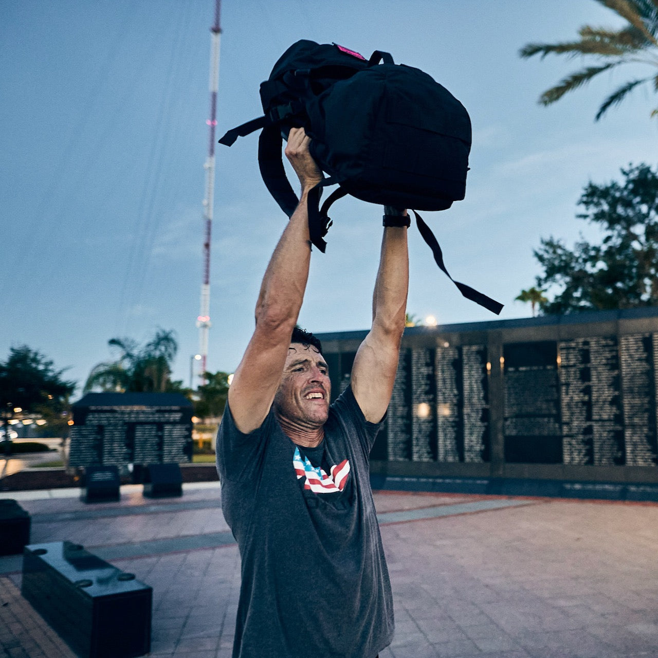 A man wearing the GORUCK Flag Spearhead Tee - Unisex Tri-BlendX in gray lifts a black backpack overhead outside by a memorial wall and palm trees, embodying the spirit of Special Forces with the iconic spearhead logo proudly displayed.
