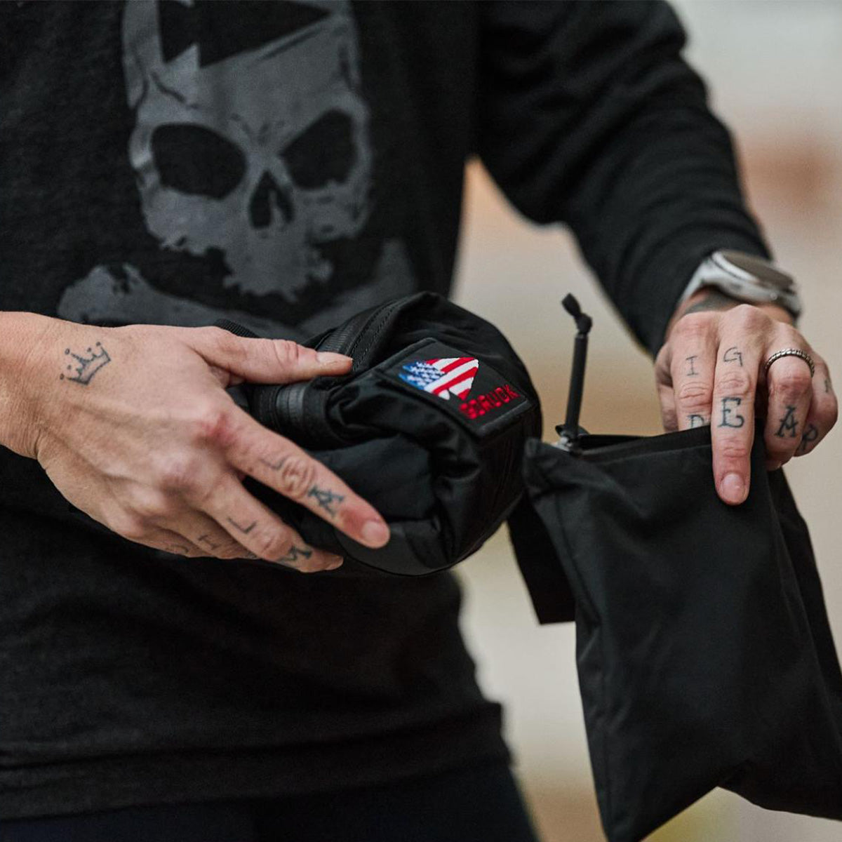 Person with tattoos on their fingers holds a black pouch with an American flag patch, wearing a skull shirt.