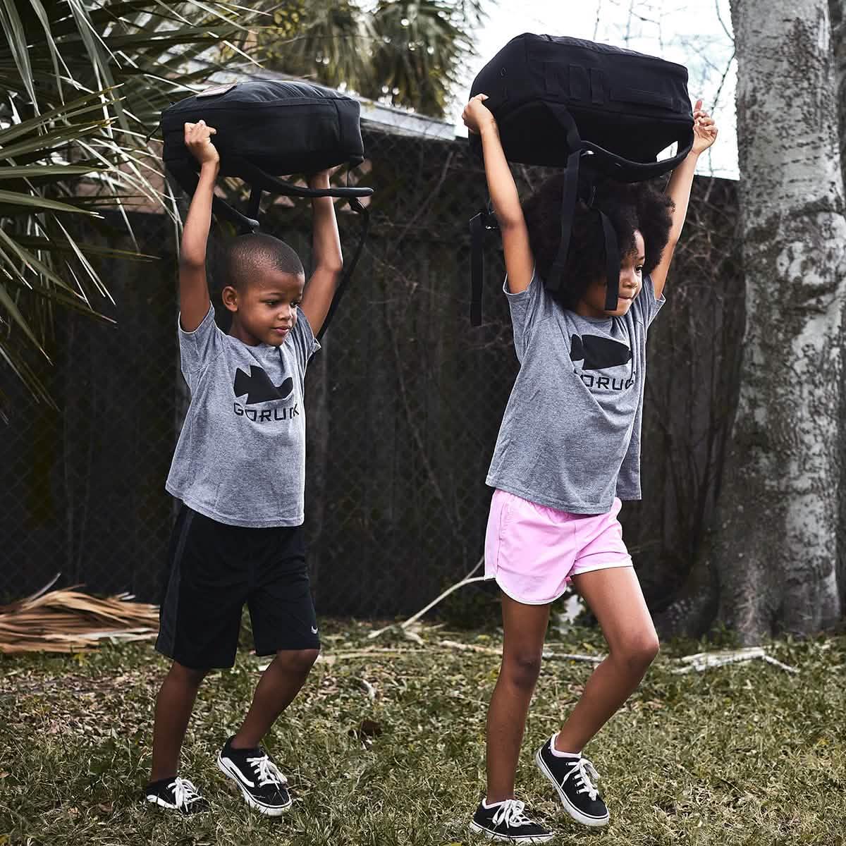 Two children in gray GORUCK t-shirts lifting black rucking bags outdoors during training
