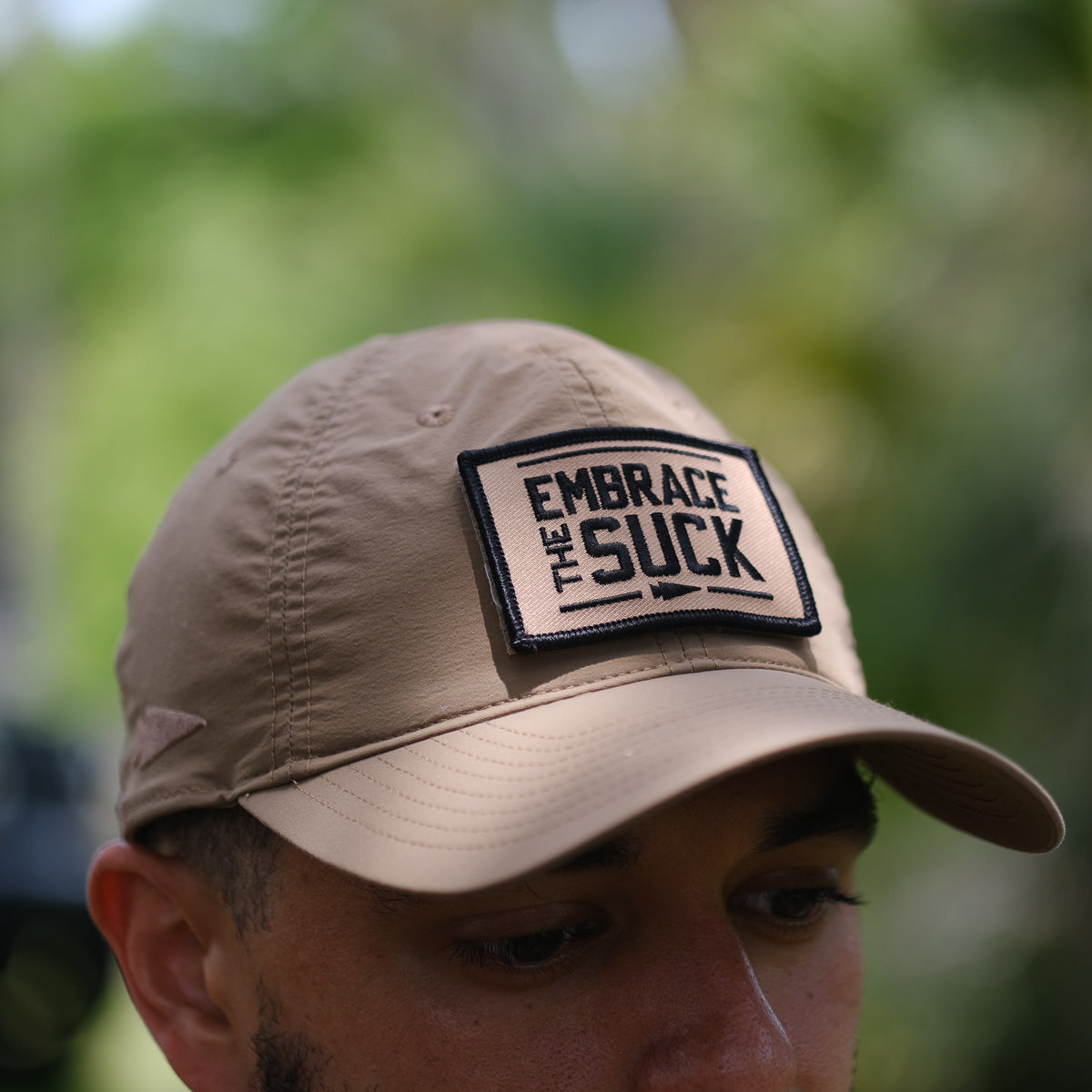 A person outdoors wears a tan cap with the Patch - Embrace The Suck, featuring an embroidered patch detail and VELCRO hook backing.