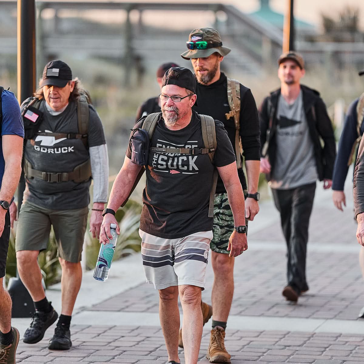A group of men with backpacks walk together on a paved path, smiling and talking with camaraderie, all wearing the Embrace the Suck Tee - Tri-Blend.