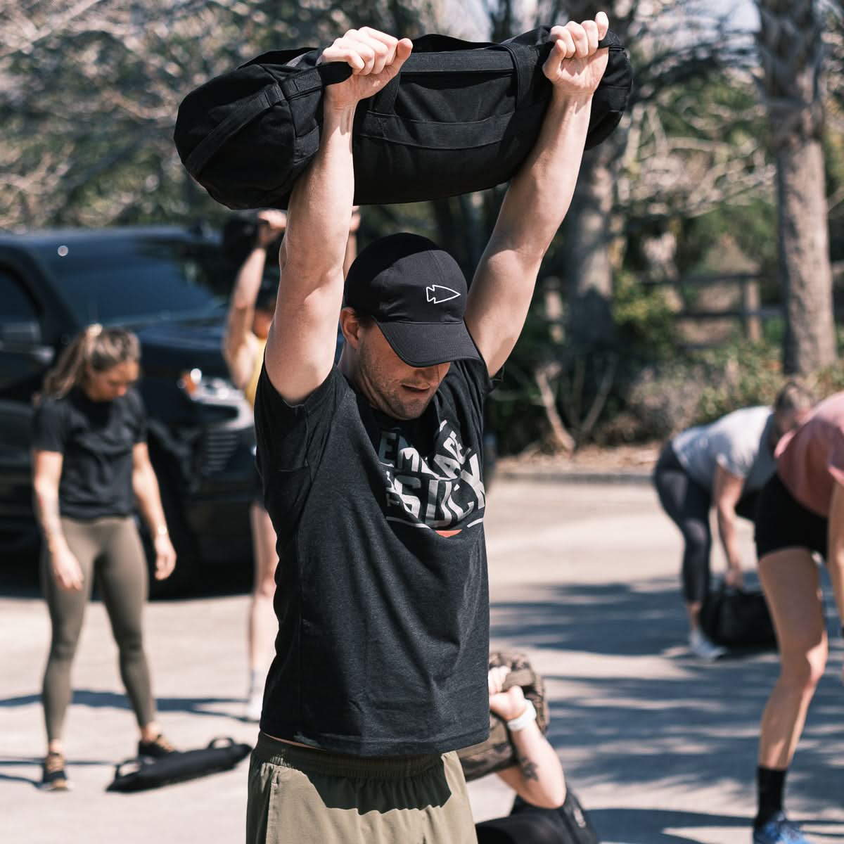 Man lifts sandbag overhead outdoors in a group workout, embodying the USMC spirit in the Embrace the Suck Tee - Tri-Blend; others exercise behind him.