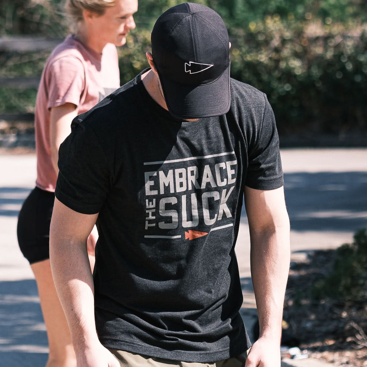 A man outdoors wears the Embrace the Suck Tee - Tri-Blend and a hat, with another person in the background—proudly displaying one of the USMC’s official mottos.