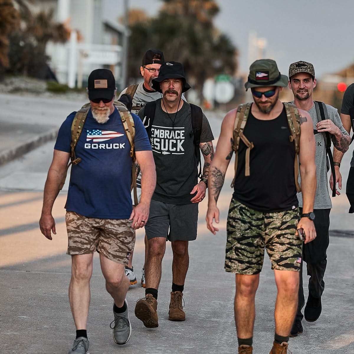 Five men in backpacks and casual athletic gear walk together outdoors, embodying the USMC spirit in Embrace the Suck Tee - Tri-Blend as they tackle challenges side by side.