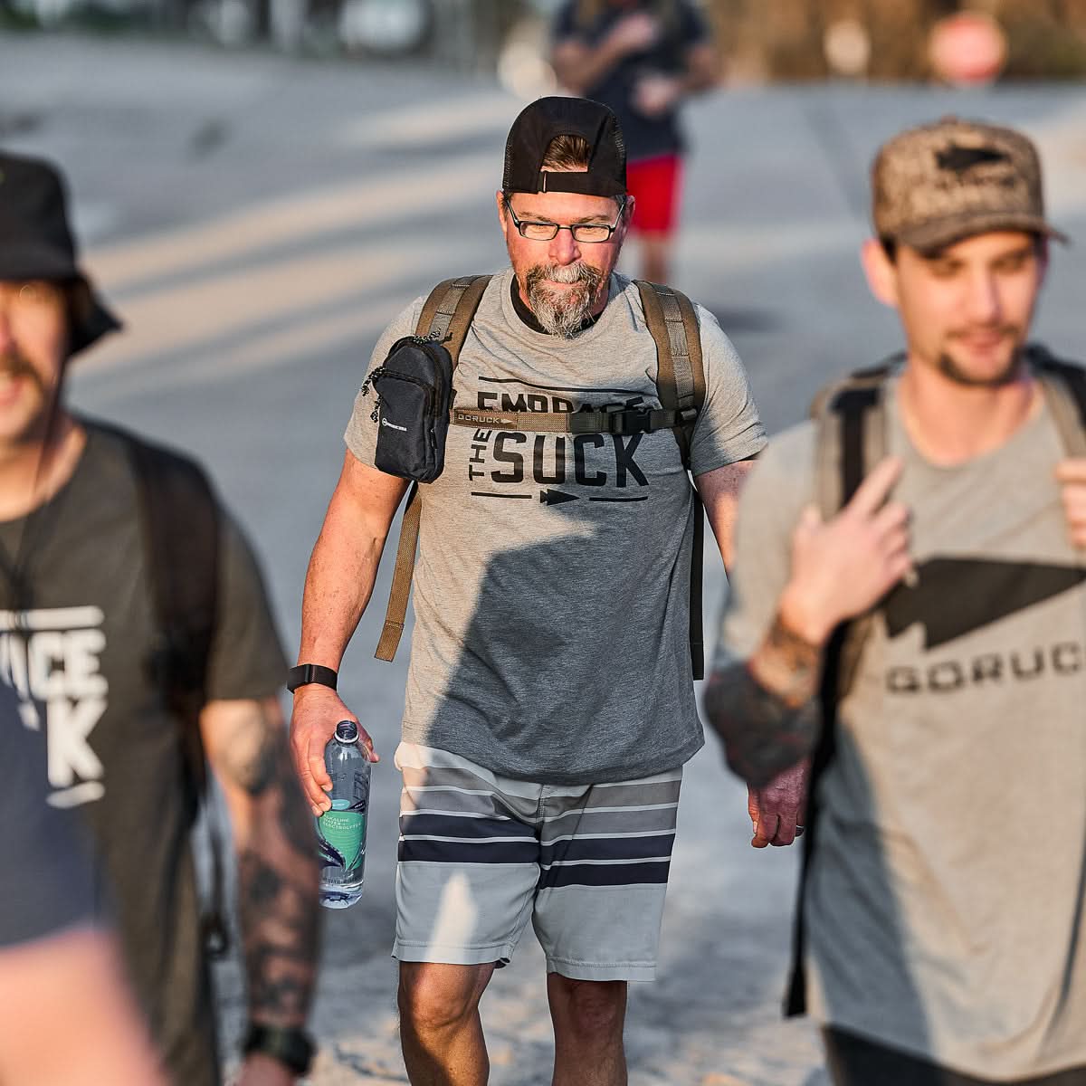 Three men in backpacks and casual clothes walk outdoors on a sunny day, ready to face challenges in the Embrace the Suck Tee - Tri-Blend.