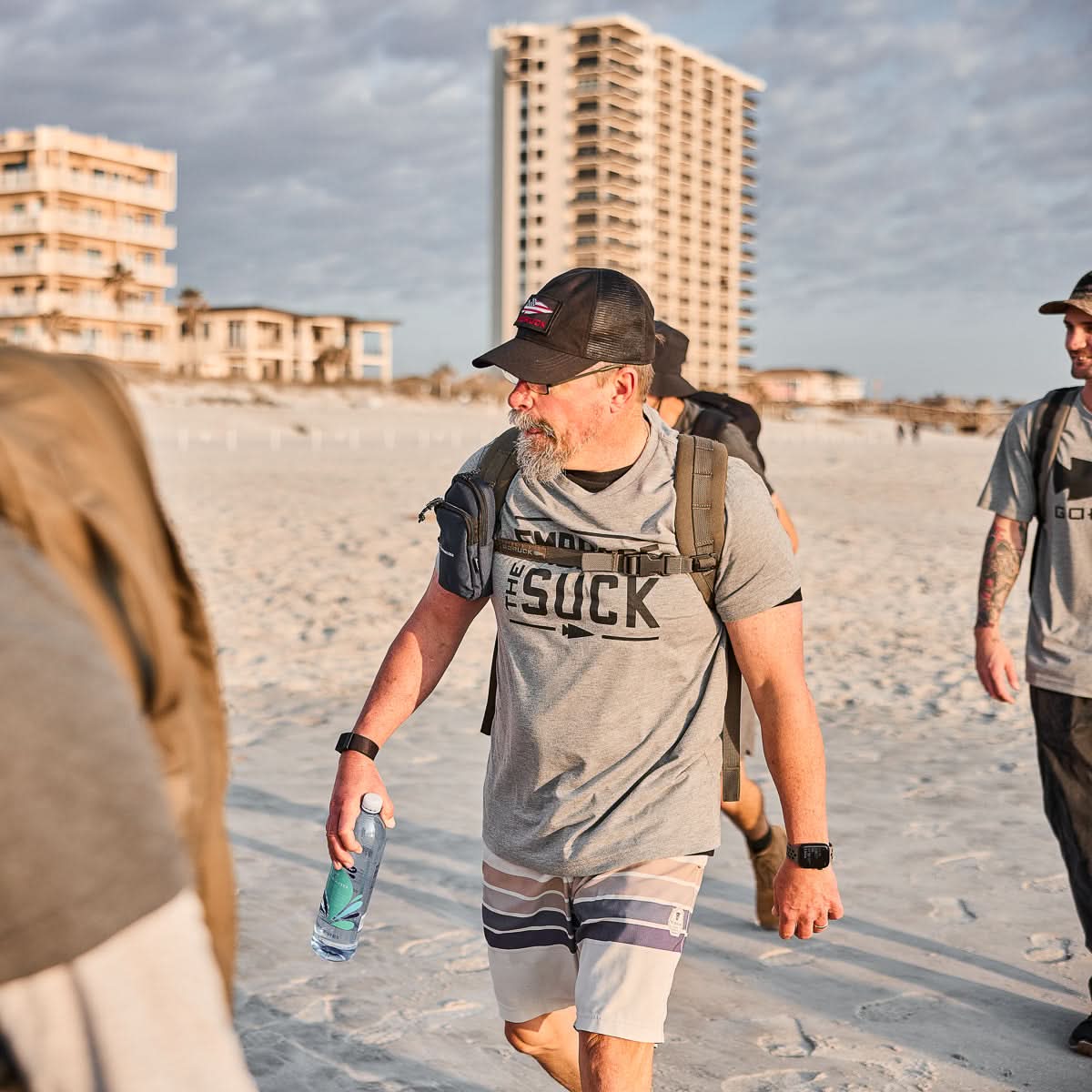 Wearing the Embrace the Suck Tee - Tri-Blend, a man in a cap and sunglasses walks on a sandy beach with a backpack and water bottle—ready to tackle any challenge, just like the USMC spirit.