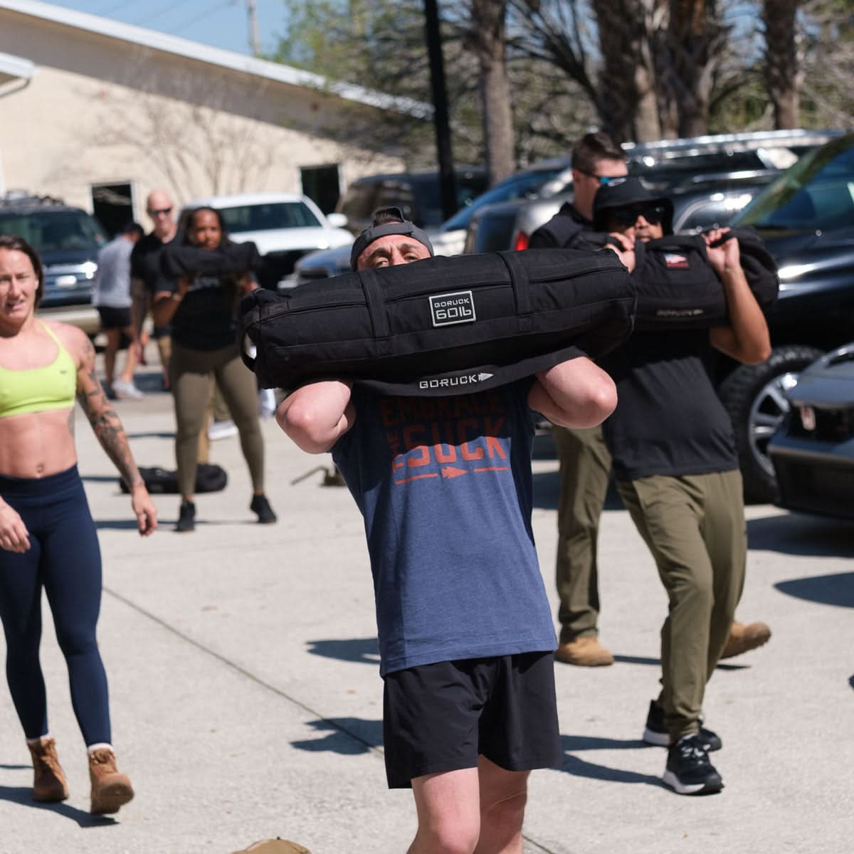 Wearing the Embrace the Suck Tee - Tri-Blend, people pushing themselves outdoors with weighted sandbags show true USMC determination and grit against a backdrop of cars and trees.