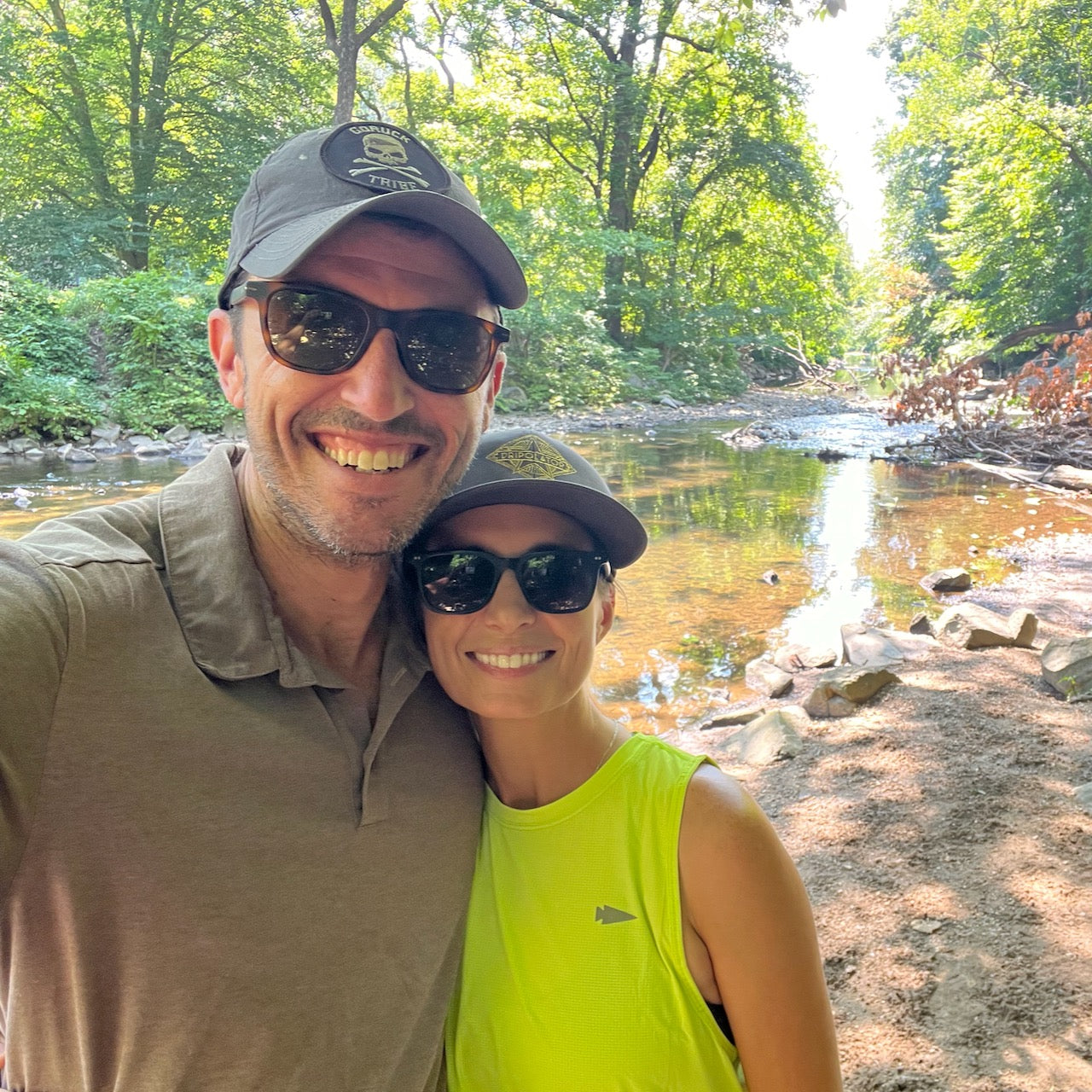 Smiling couple in sunglasses and hats poses by a sunlit creek in a leafy forest, with her wearing the Women’s USA Performance Tank made from breathable Featherweight ToughMesh fabric.