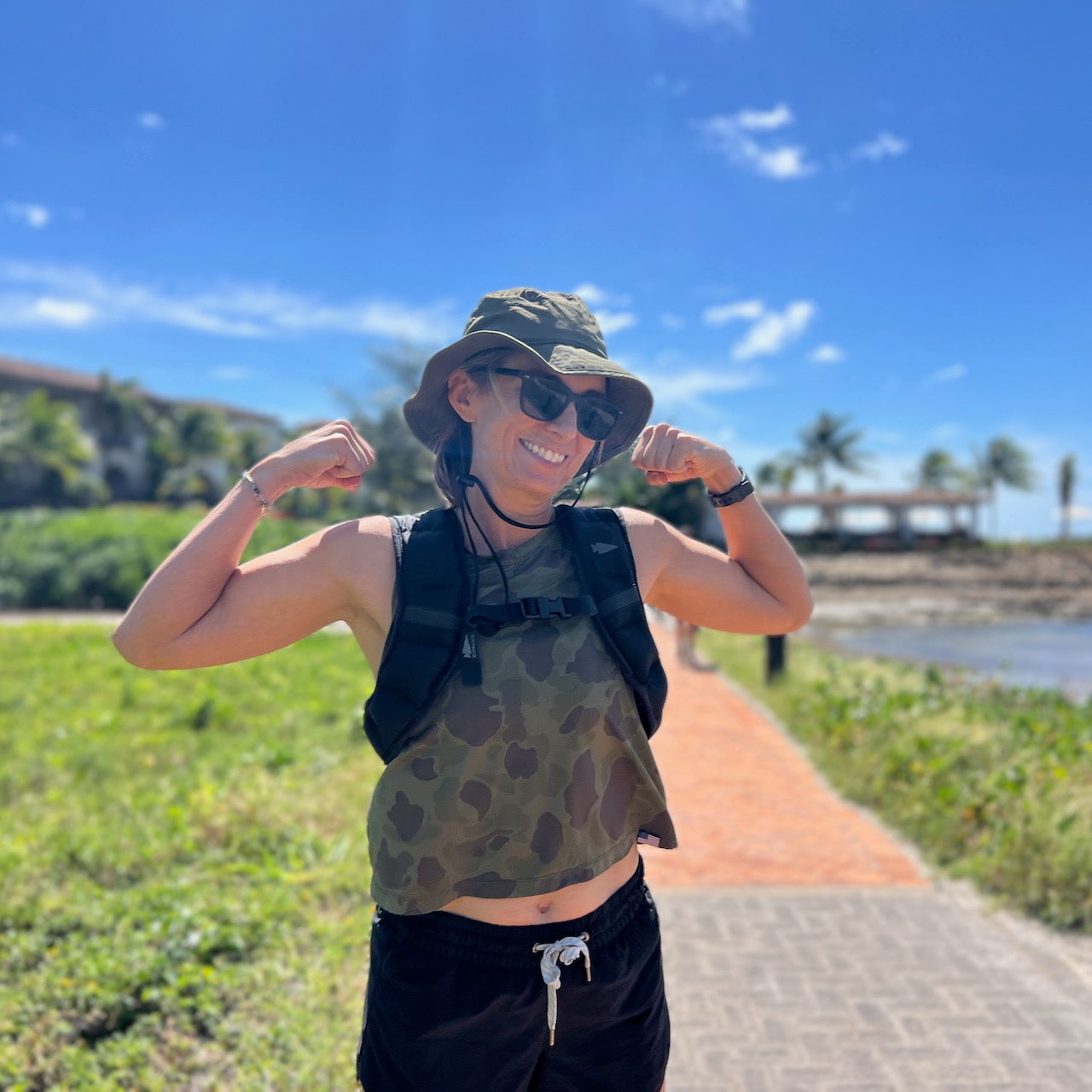 Smiling woman in sunglasses and a hat poses outdoors near the beach on a sunny day, wearing the Spy Ruck | Women's Weighted Vest.