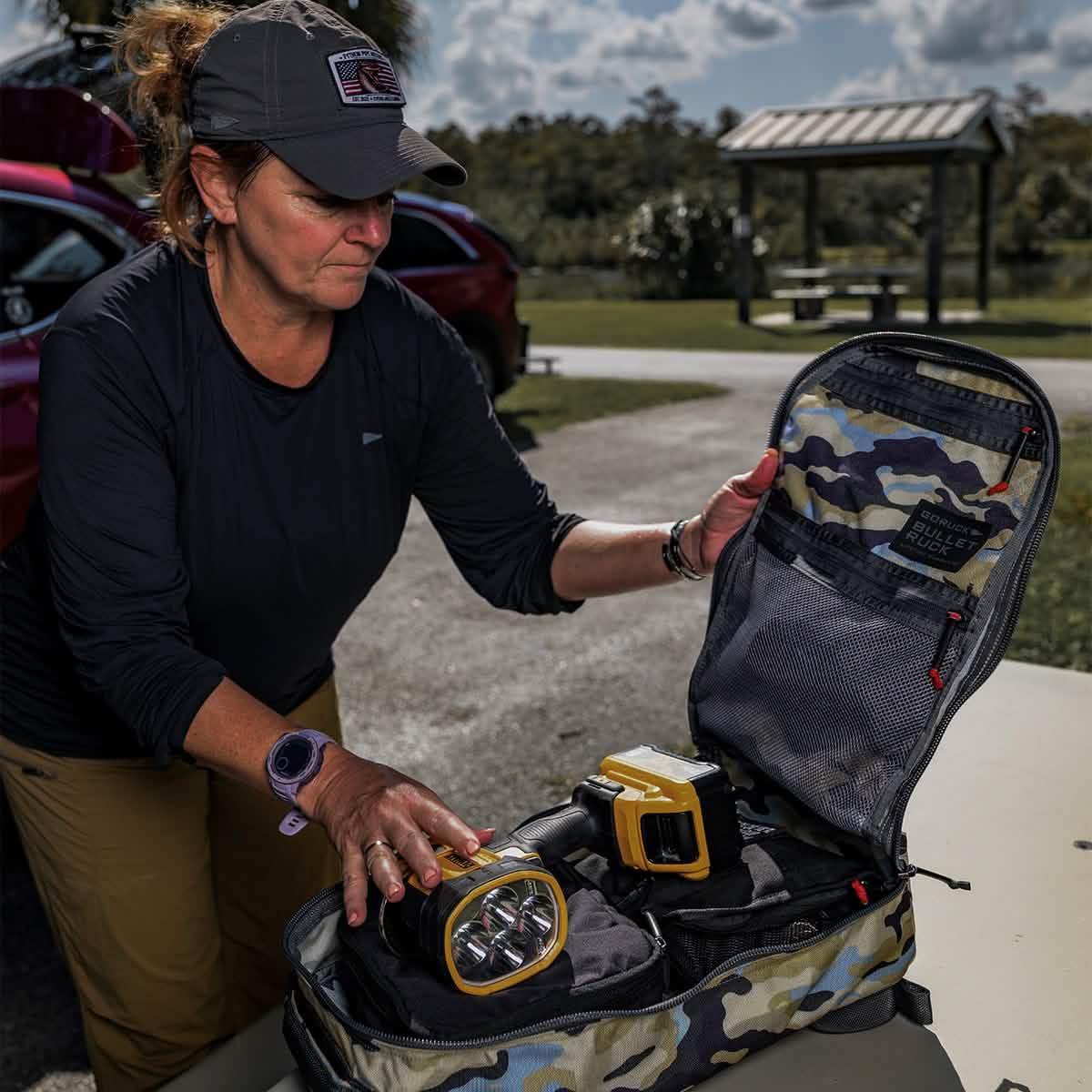 Woman unpacking GORUCK Everglades Camo Bullet ruck case with yellow flashlights on outdoor table