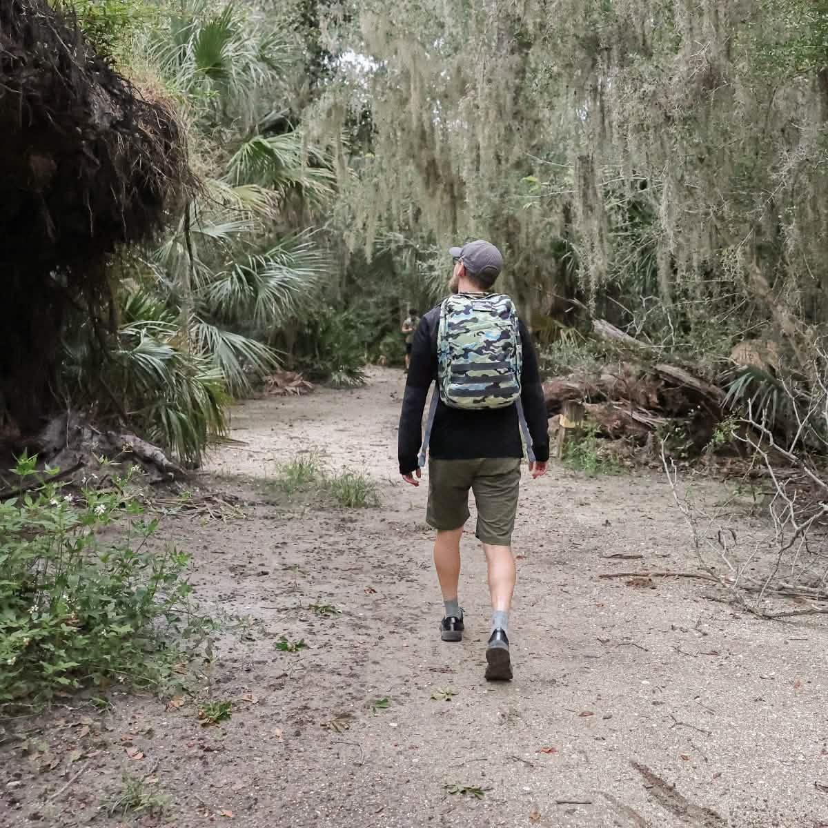 Man wearing Everglades camo backpack hiking on wooded trail with dense green foliage
