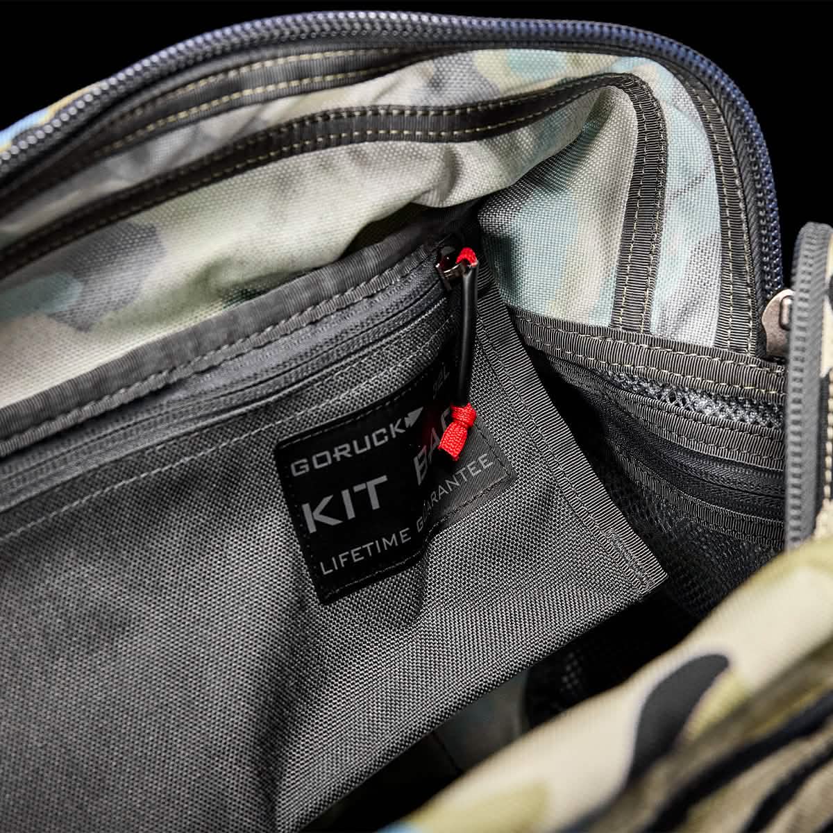 Close-up of the inside of an open camouflage Kit Bag by GORUCK, with a stitched label reading GORUCK KIT. A red zipper pull suggests inner compartments, and the interior pattern matches the exterior design.