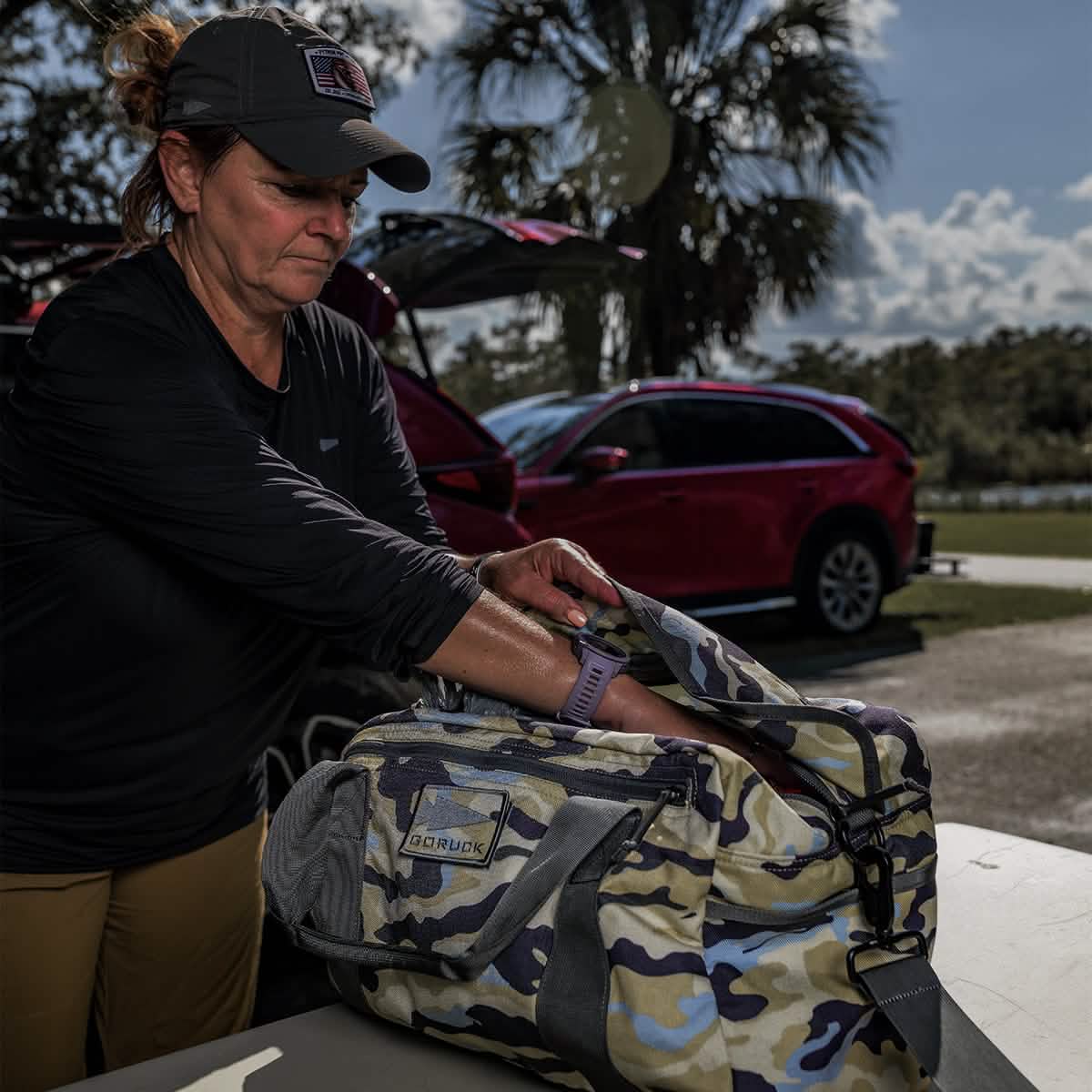 A woman wearing a cap and long-sleeved shirt is packing items into a GORUCK Kit Bag (Includes Shoulder Strap) on a table. A red SUV with an open trunk stands in the background, surrounded by trees under a sunny sky.